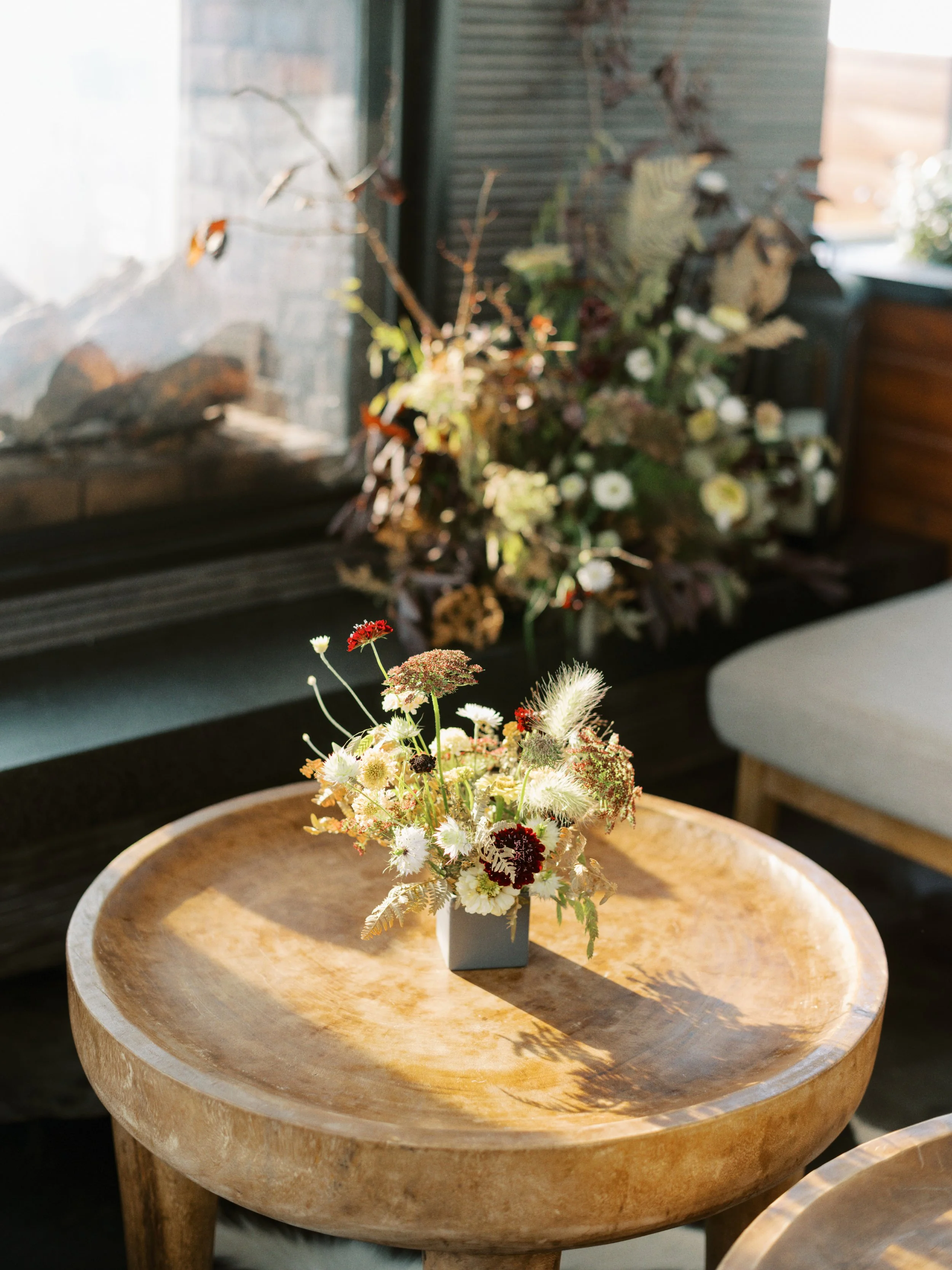 A wooden side table with a small floral arrangement in a gray square vase, featuring various small white, red, and purple flowers and greenery.