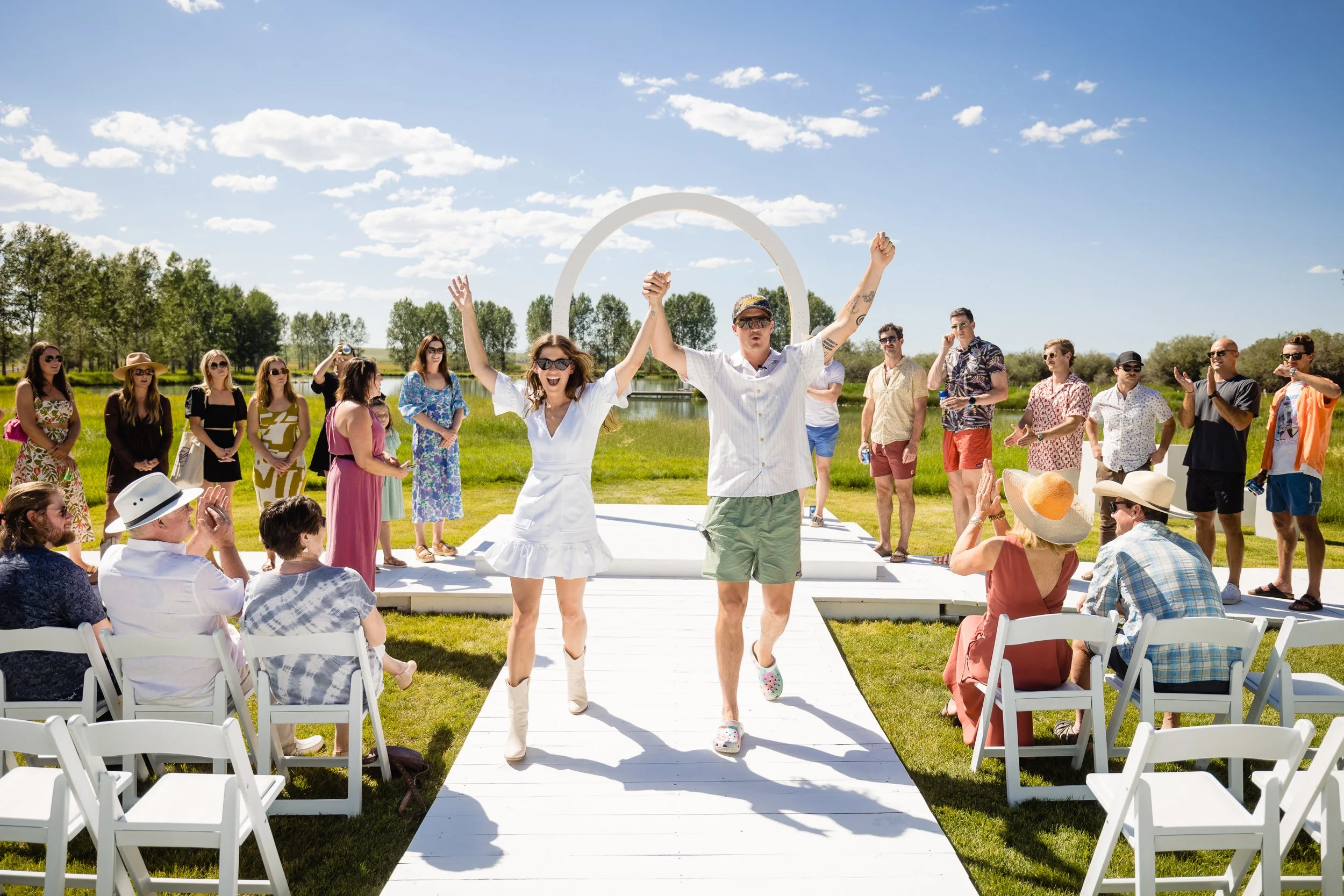 Couple walking down an aisle outdoors on a sunny day, with friends and family clapping and watching. The backdrop includes trees, a lake, and a blue sky with clouds.