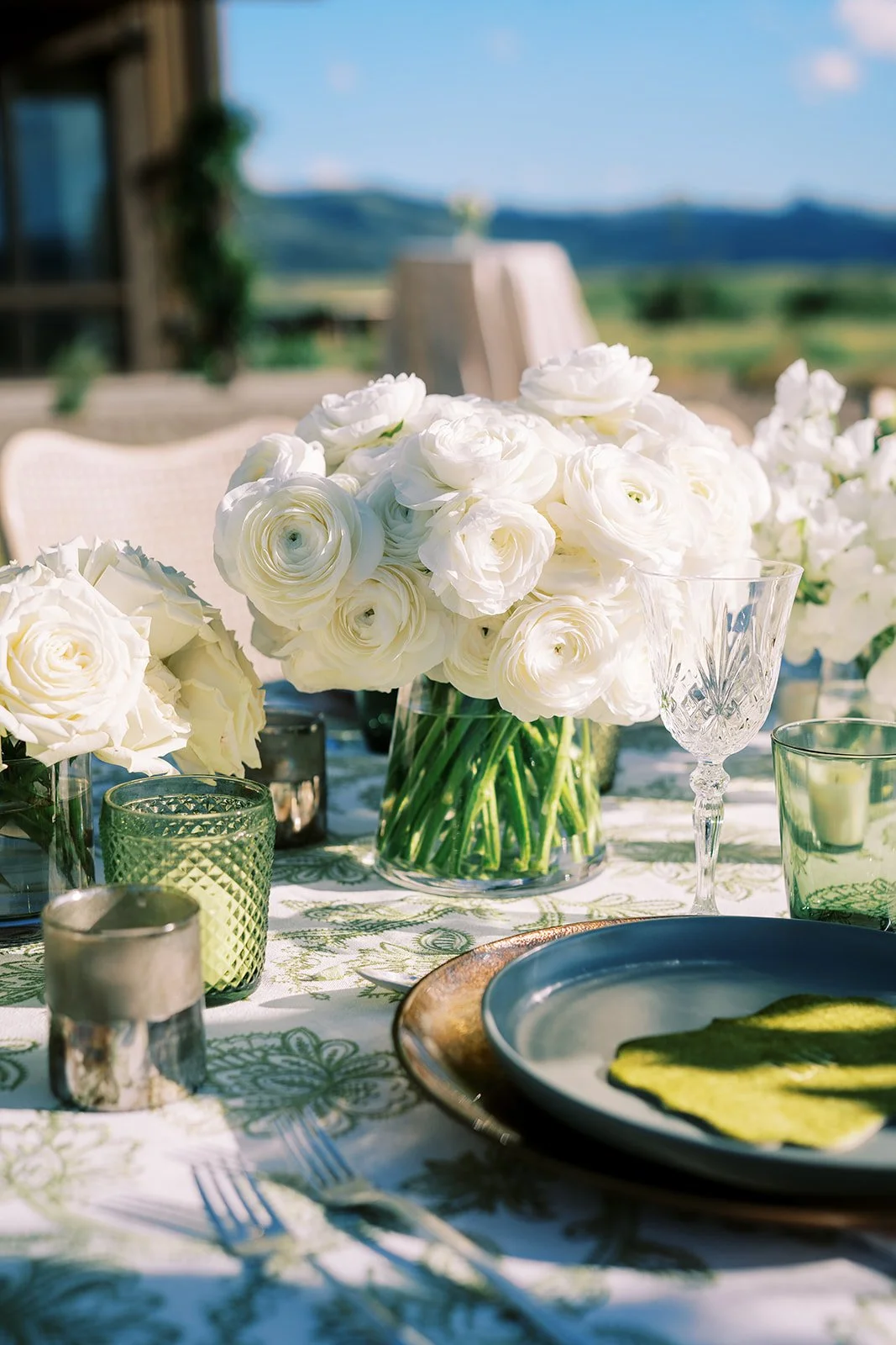 Table setting with white floral centerpiece, glassware, and decorative candles outdoors with scenic landscape in the background.