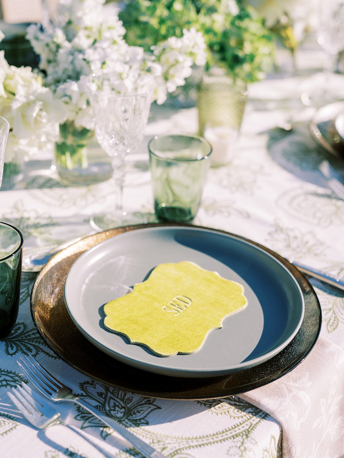 A table set for an outdoor meal with white flowers in glass vases, green-tinted glasses, and a plate with a yellow paper napkin that has a cutout of the state of Wisconsin and the word "SIED" in the center.