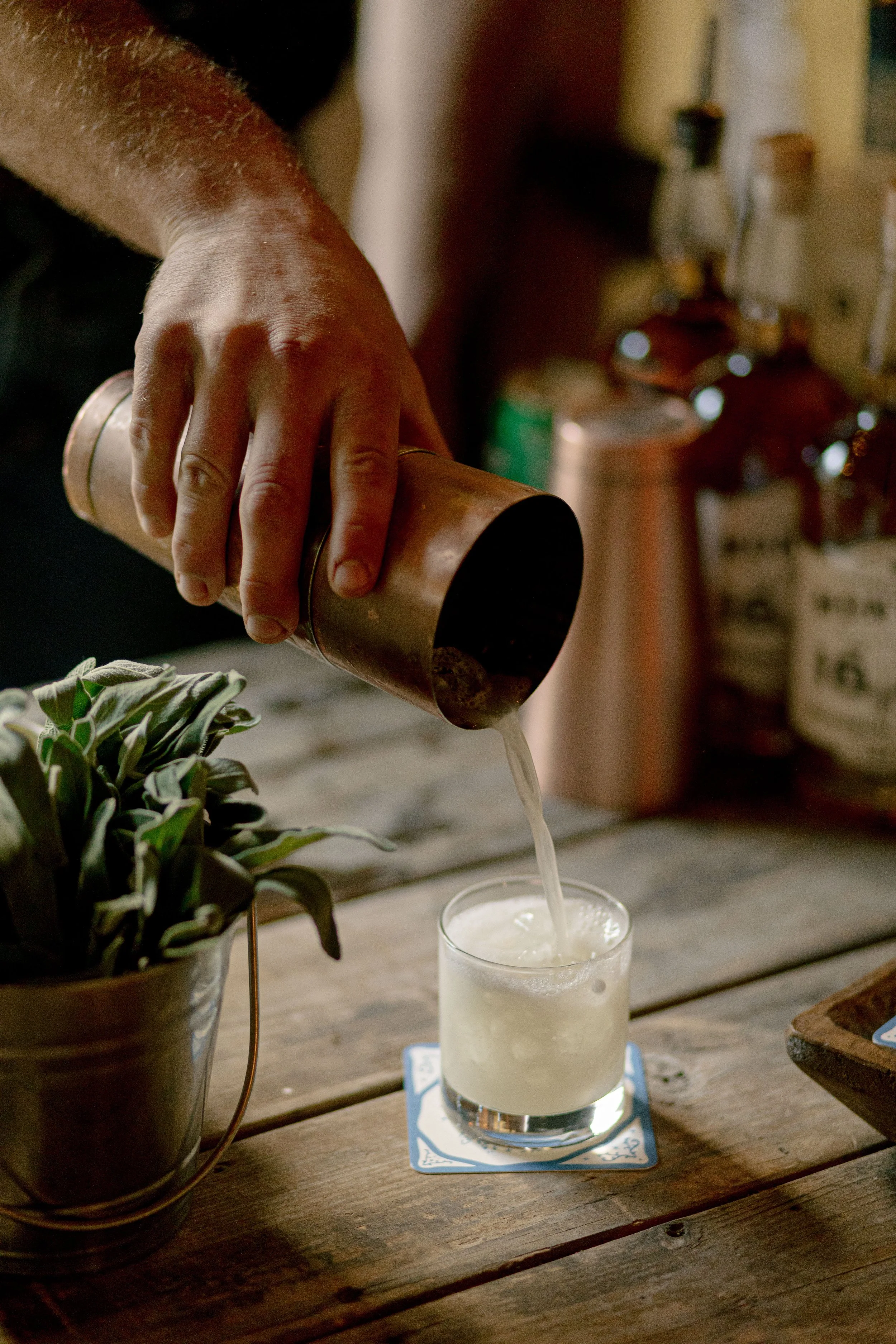 A person is pouring a beverage from a copper mug into a glass on a wooden table. There is a potted plant with dark green leaves nearby, and the background is slightly blurred with various jars and bottles.