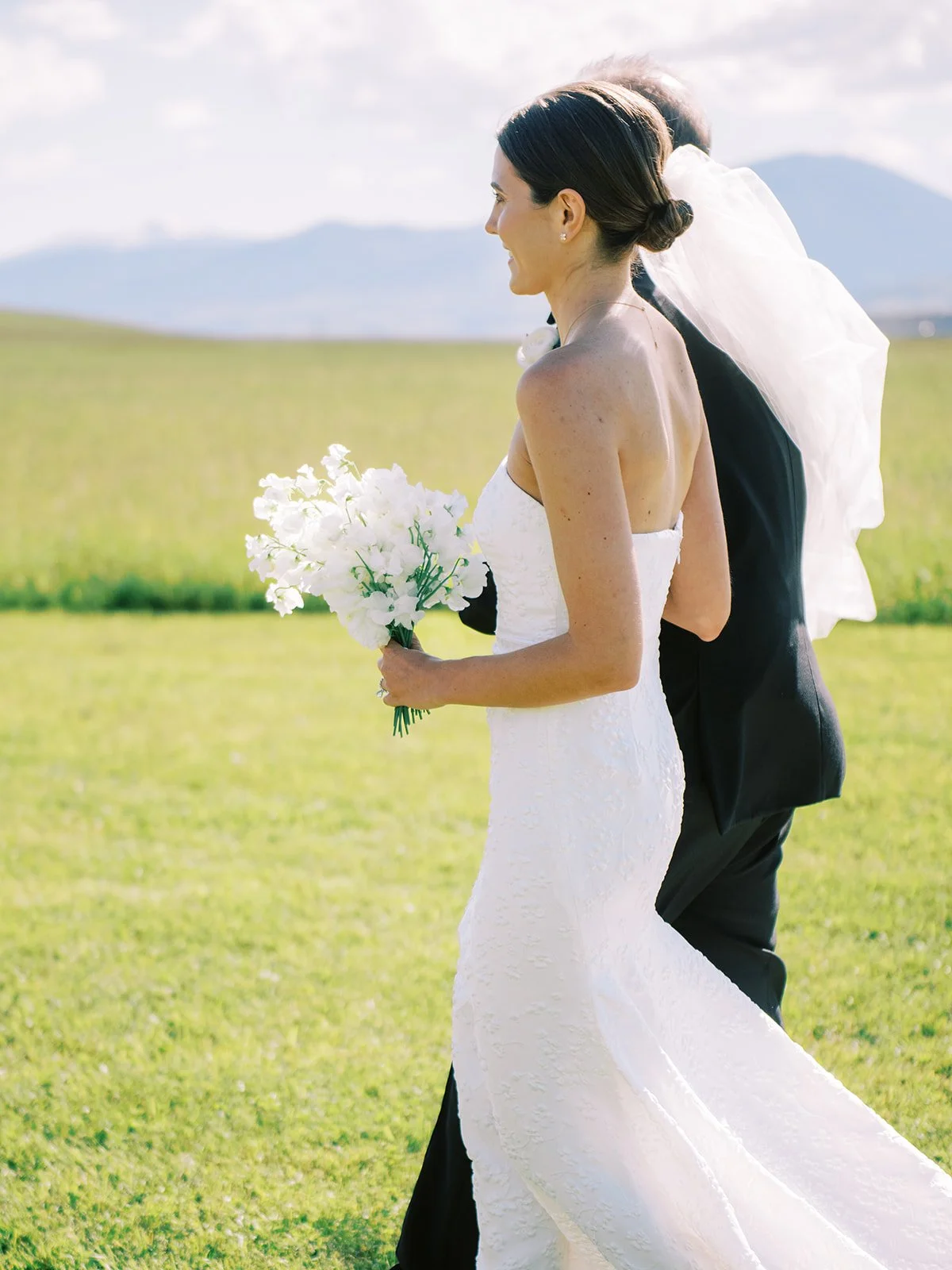 A bride in a white wedding dress holding a bouquet of white flowers, standing outdoors on a grassy field with mountains in the background.