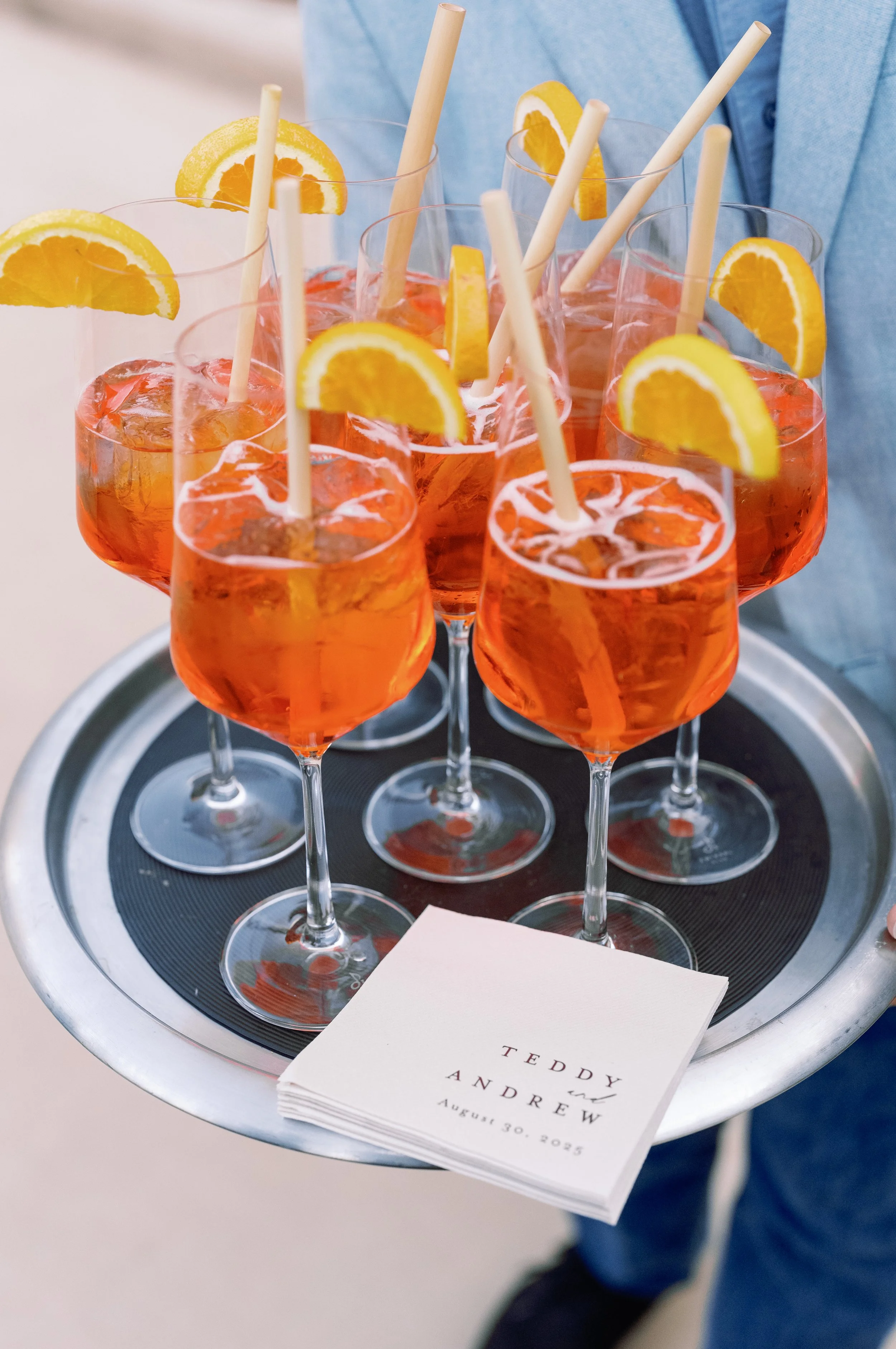 A tray holding several glasses of orange-colored drinks garnished with orange slices and straws, with a wedding program or menu placed on the tray that reads 'Teddy and Audrey August 30, 2025'.