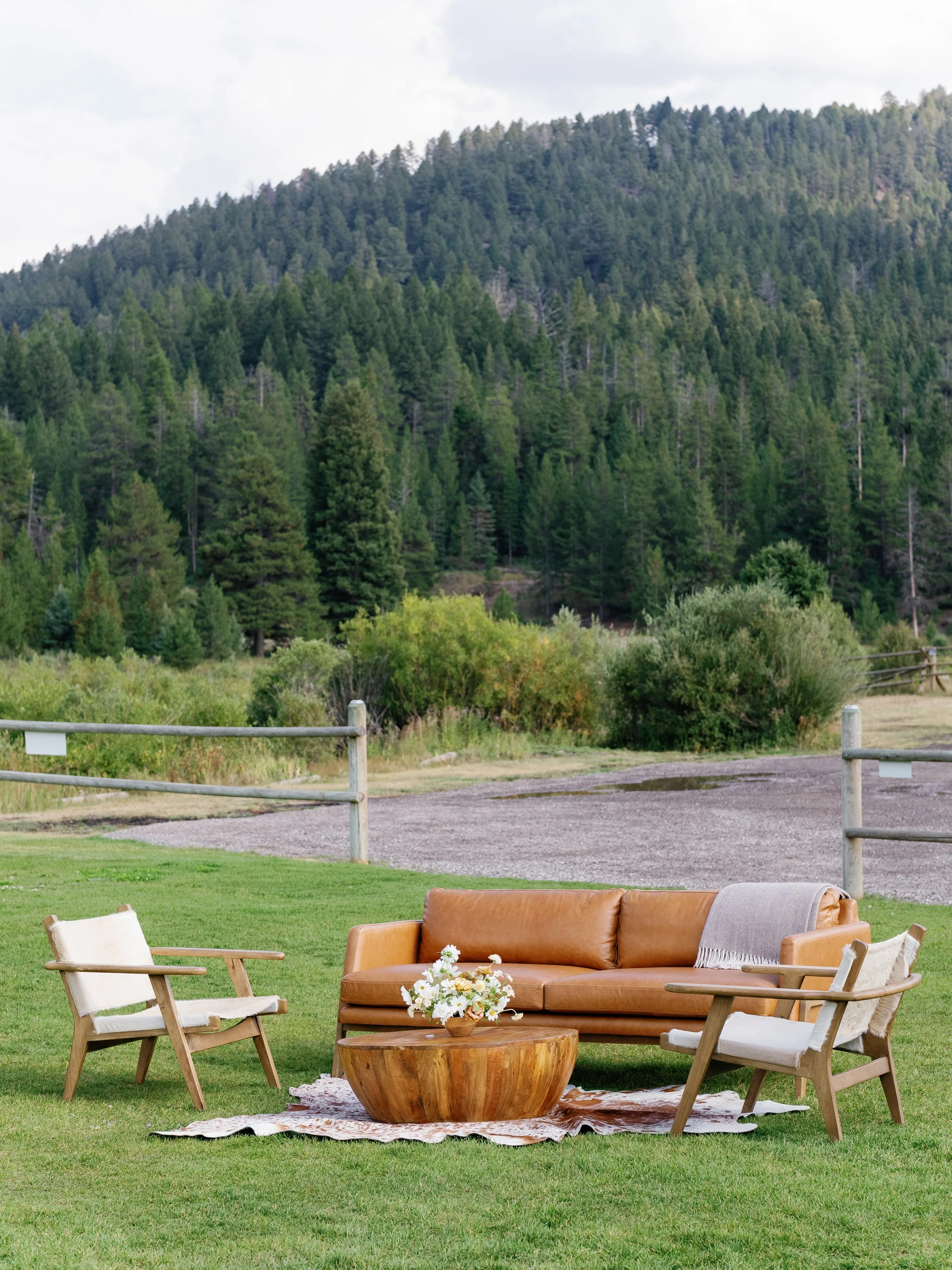 Outdoor patio with brown leather sofa, wooden chairs, and a wooden coffee table with a flower arrangement, surrounded by green grass and a backdrop of forested hills.