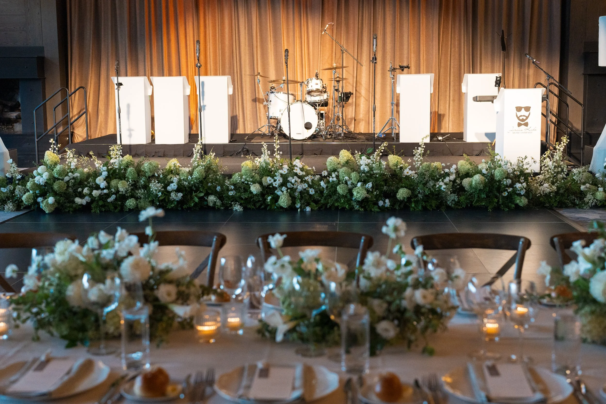 A stage set for a music performance with a drum kit and multiple microphones, decorated with flowers in front, and a golden curtain backdrop.