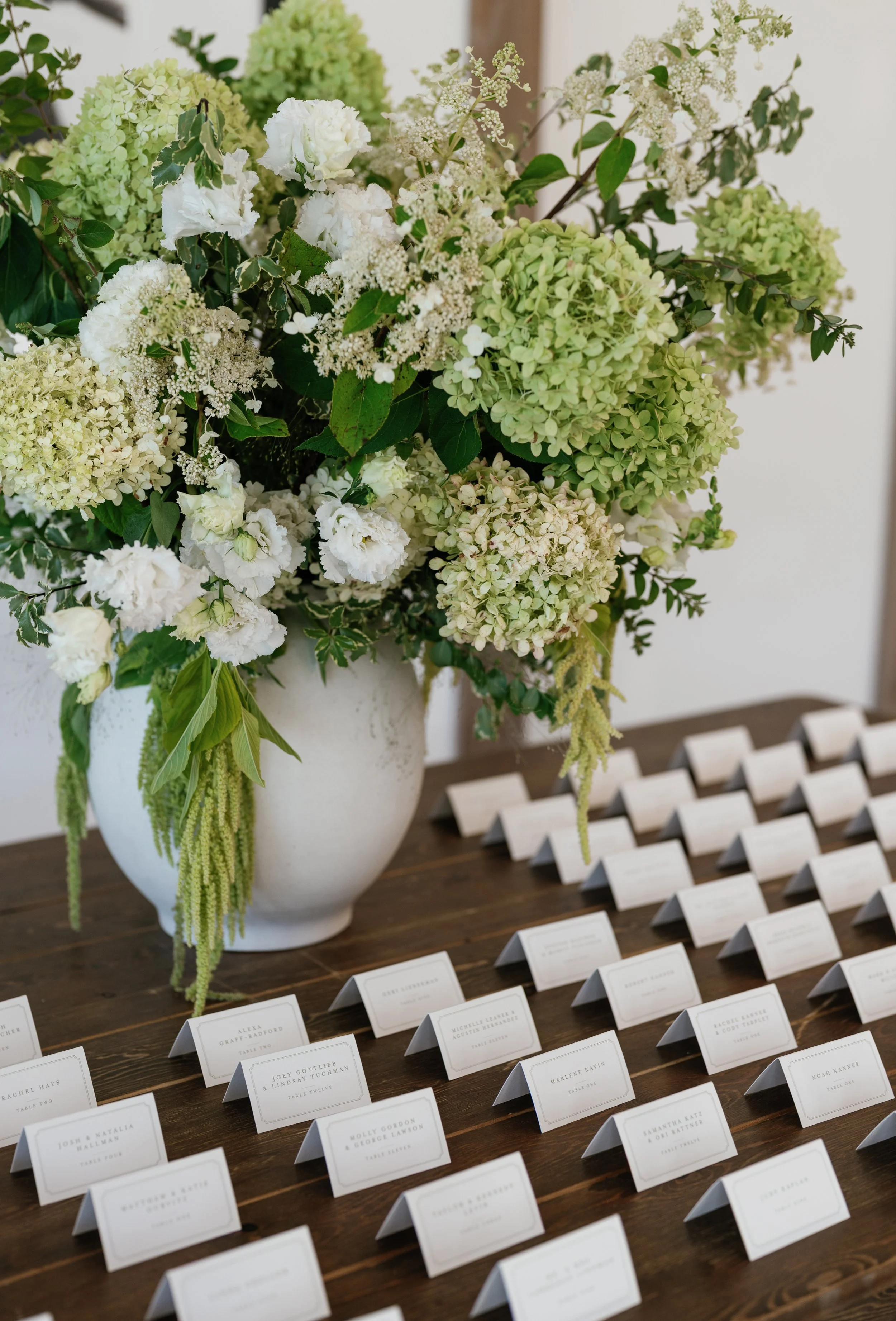 A wedding or event seating chart with name cards on a wooden table next to a large floral arrangement with white and green flowers in a white vase.