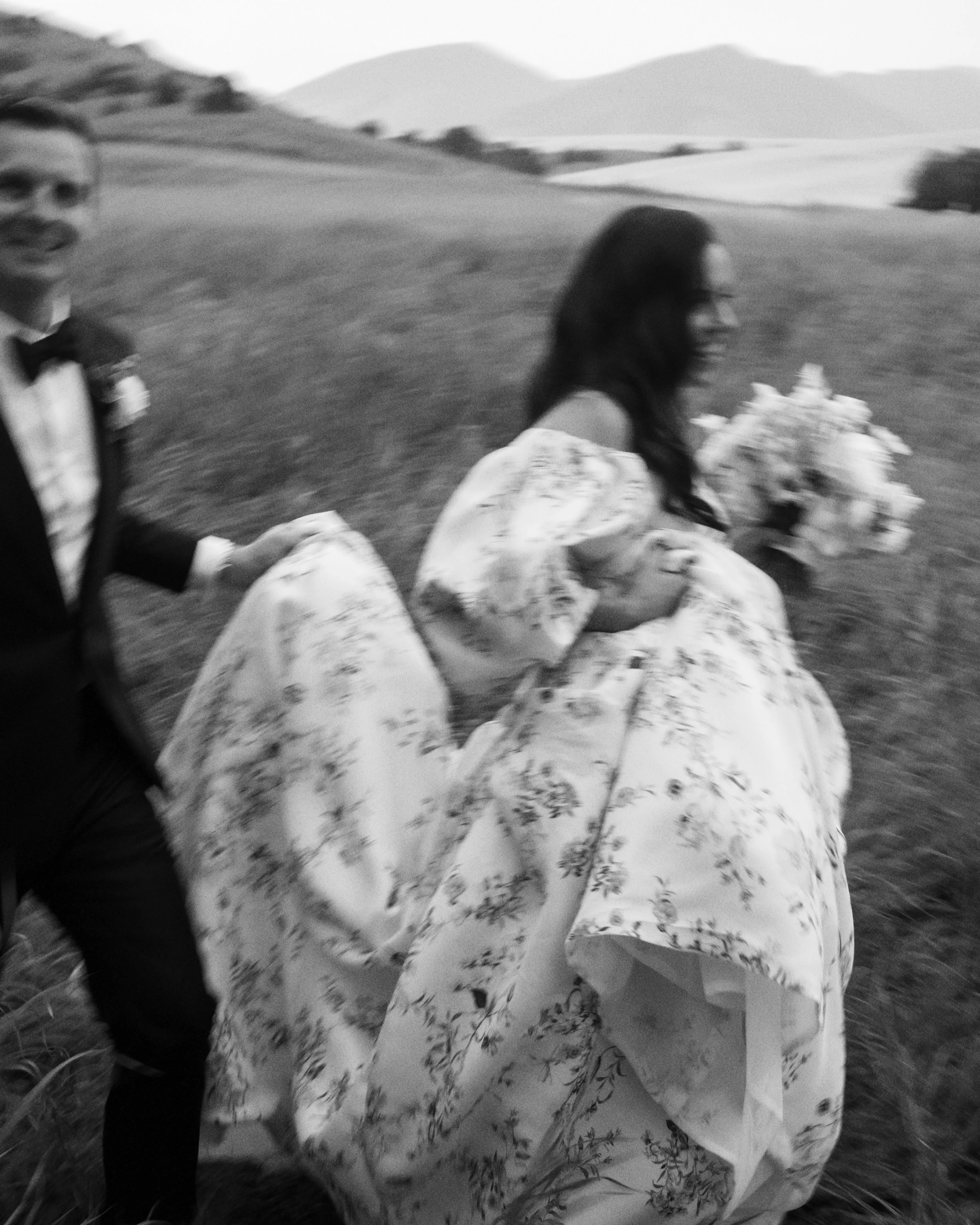Black-and-white photo of a woman in a floral dress holding hands with a man in a tuxedo, walking outdoors with hills in the background.