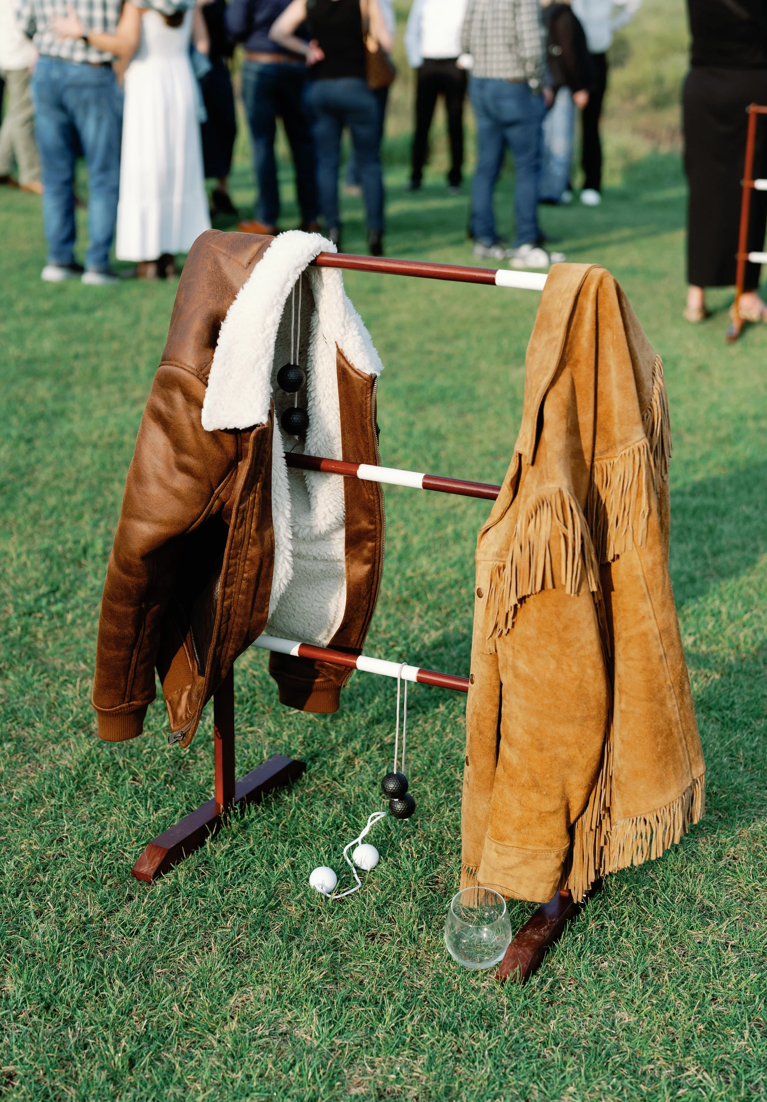 A ring toss game setup with two fringed and leather jackets hanging on a wooden frame, and three white balls on a string on the grass, with a group of people gathered in the background.