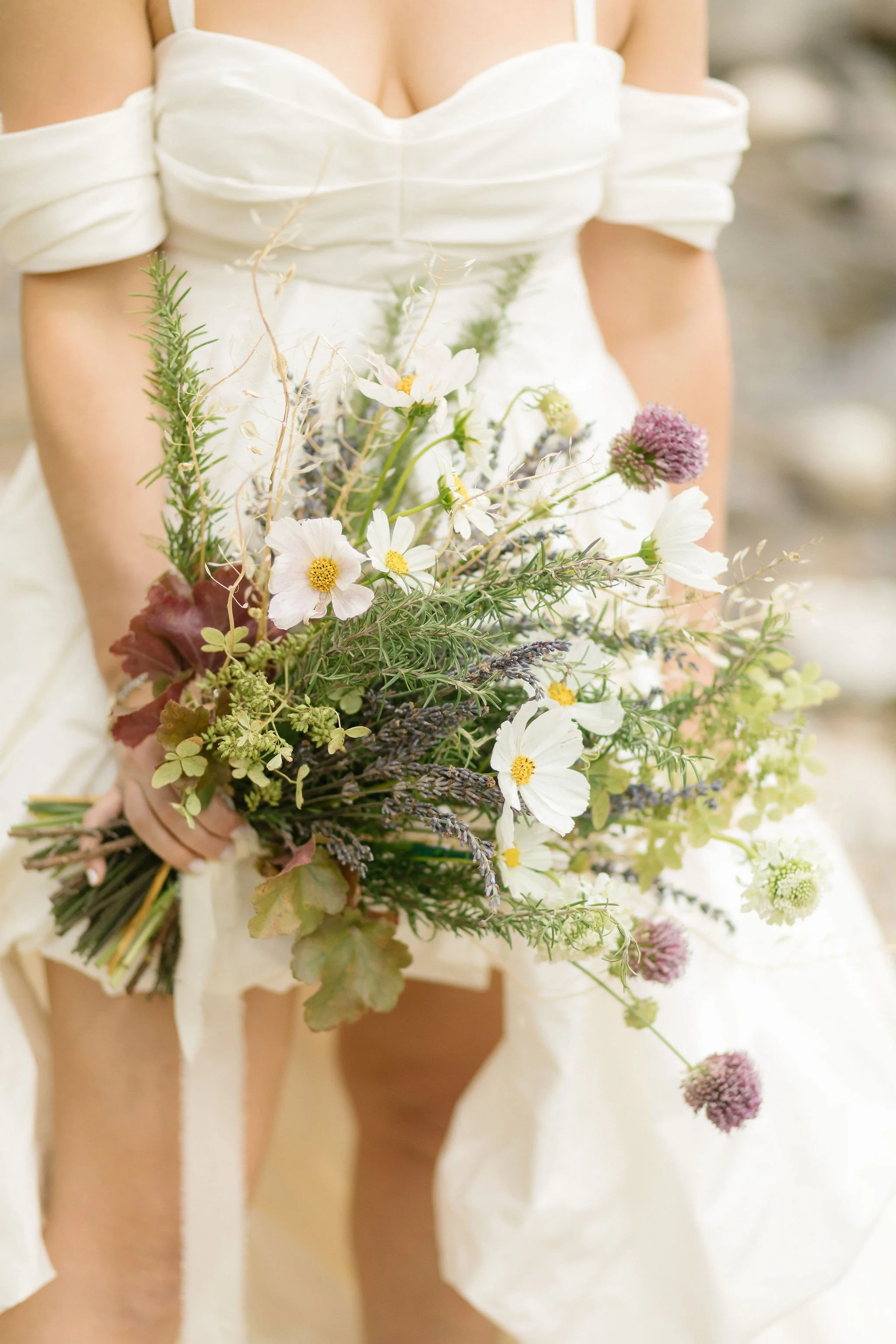 Person in a white off-shoulder dress holding a bouquet of mixed flowers and greenery.