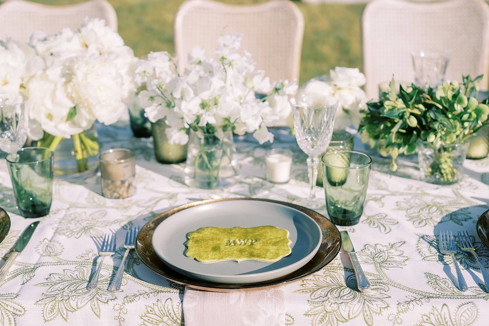 Elegant dining setup with a white floral tablecloth, green glassware, candles, and a floral centerpiece with predominantly white flowers. A plate with a yellow decorative napkin with initials is at the center.