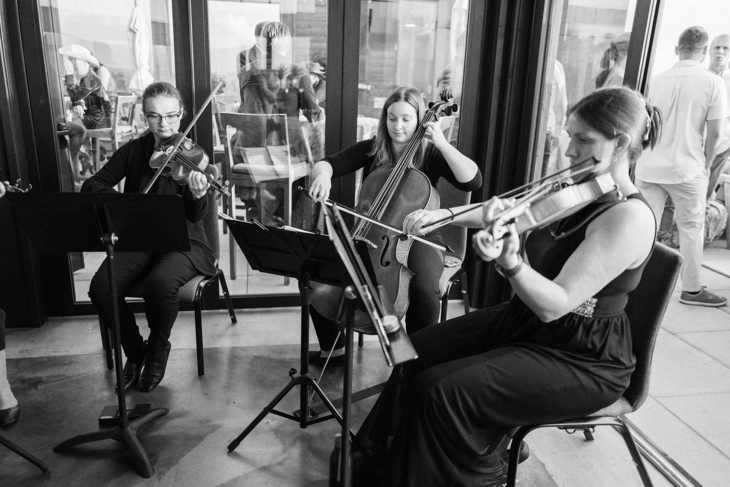 Three young women playing violins and a woman playing a cello in a room with glass walls, audience members visible through the glass, some talking and sitting.
