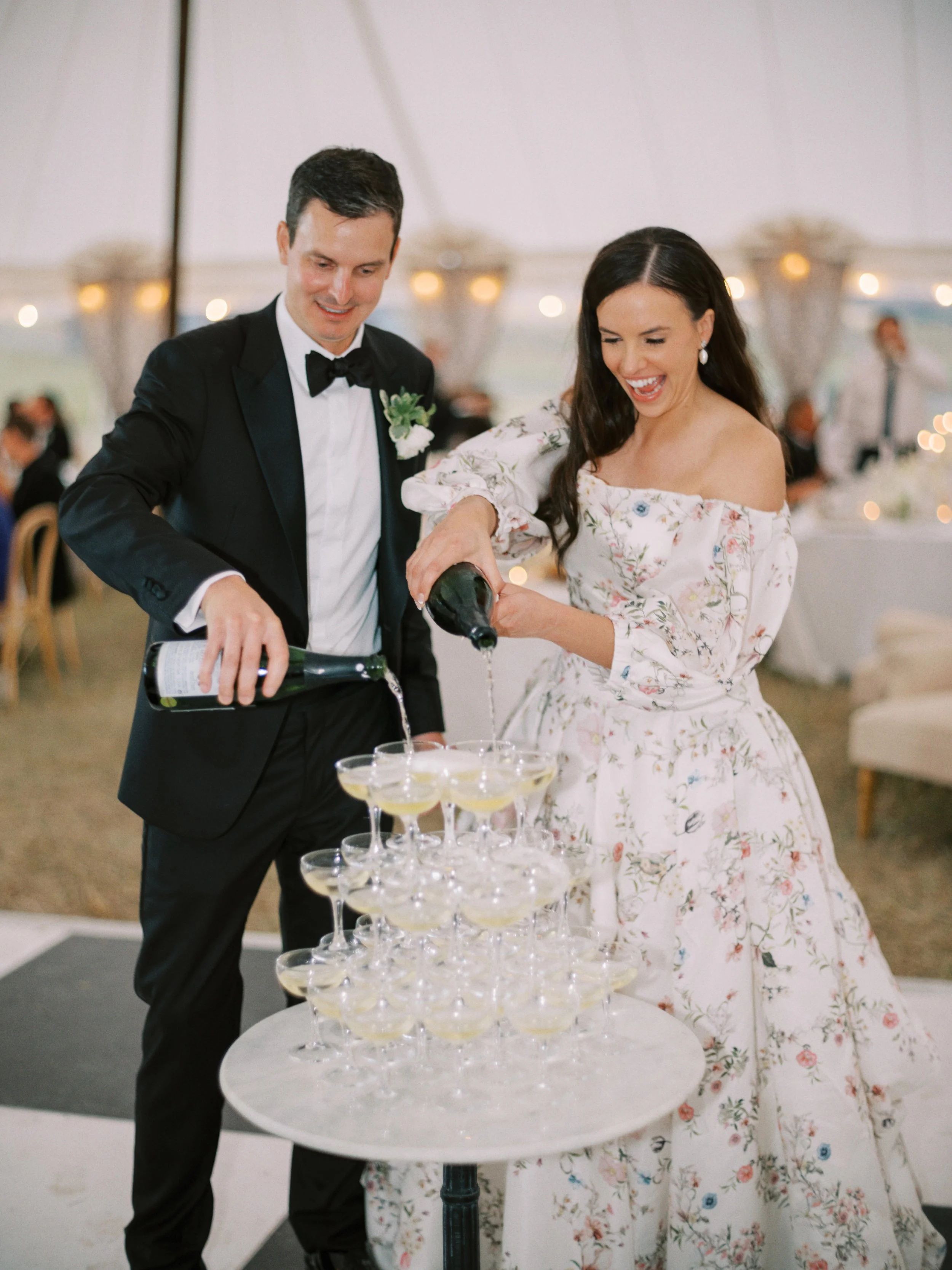 A couple in formal attire, the man in a tuxedo and the woman in a floral dress, celebrating with champagne at a wedding reception. They are pouring champagne into a pyramid of glasses.