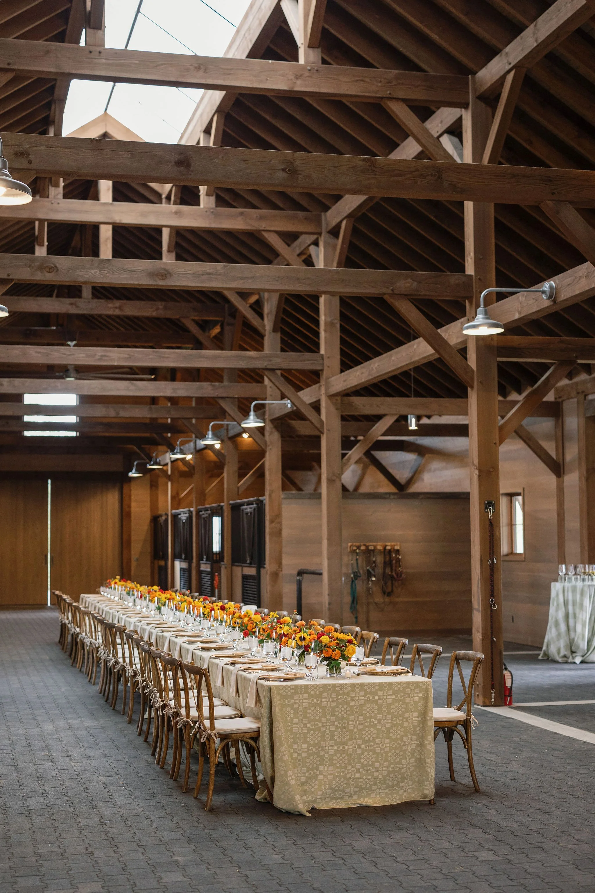 Long rustic barn reception with warm string lighting overhead, extended dining tables, and bright floral centerpieces, designed by Wild Blume