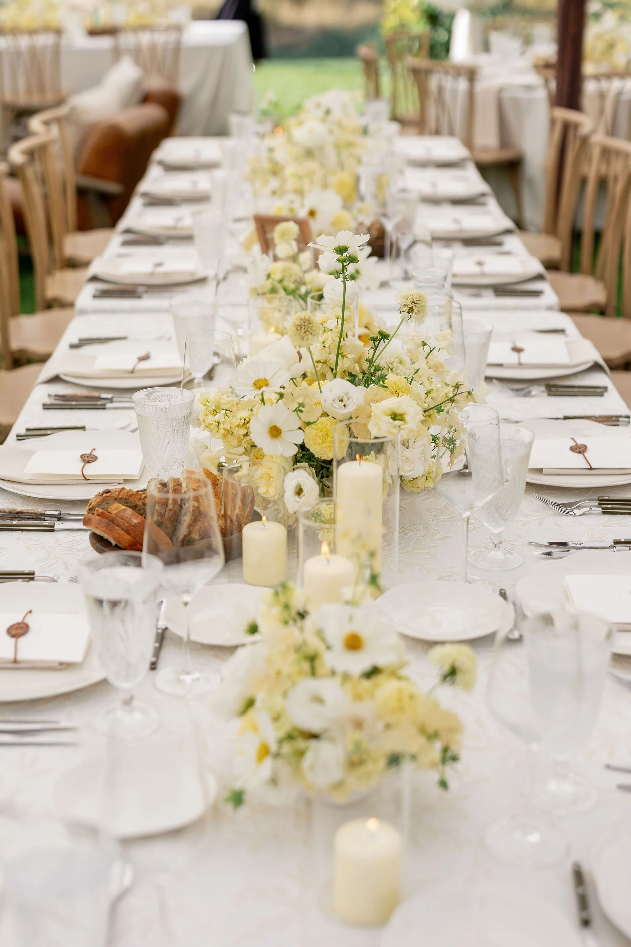 Long wedding reception table with white linen, green and white floral runners, candles, and golden glassware inside a tent, by Wild Blume