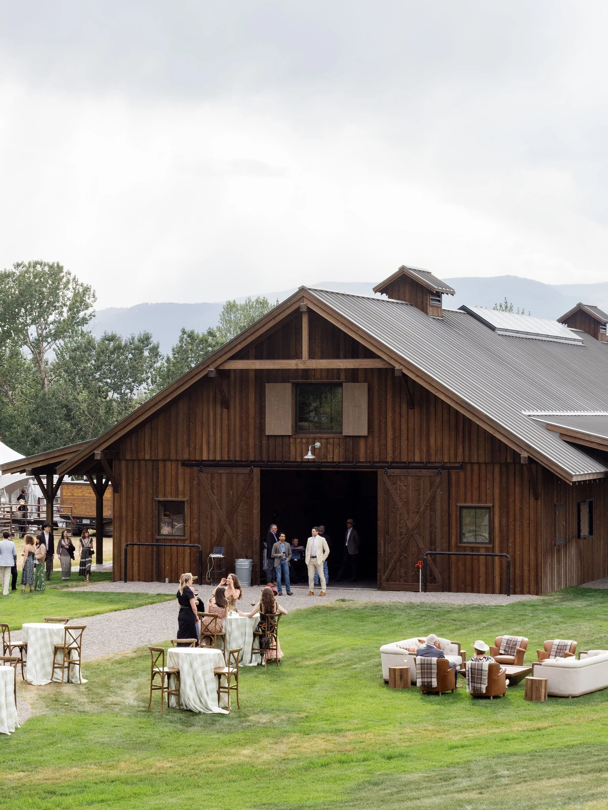 Outdoor wedding lounge setup with white sofas, leather chairs, and wood stump accents beside a rustic barn, designed by Wild Blume