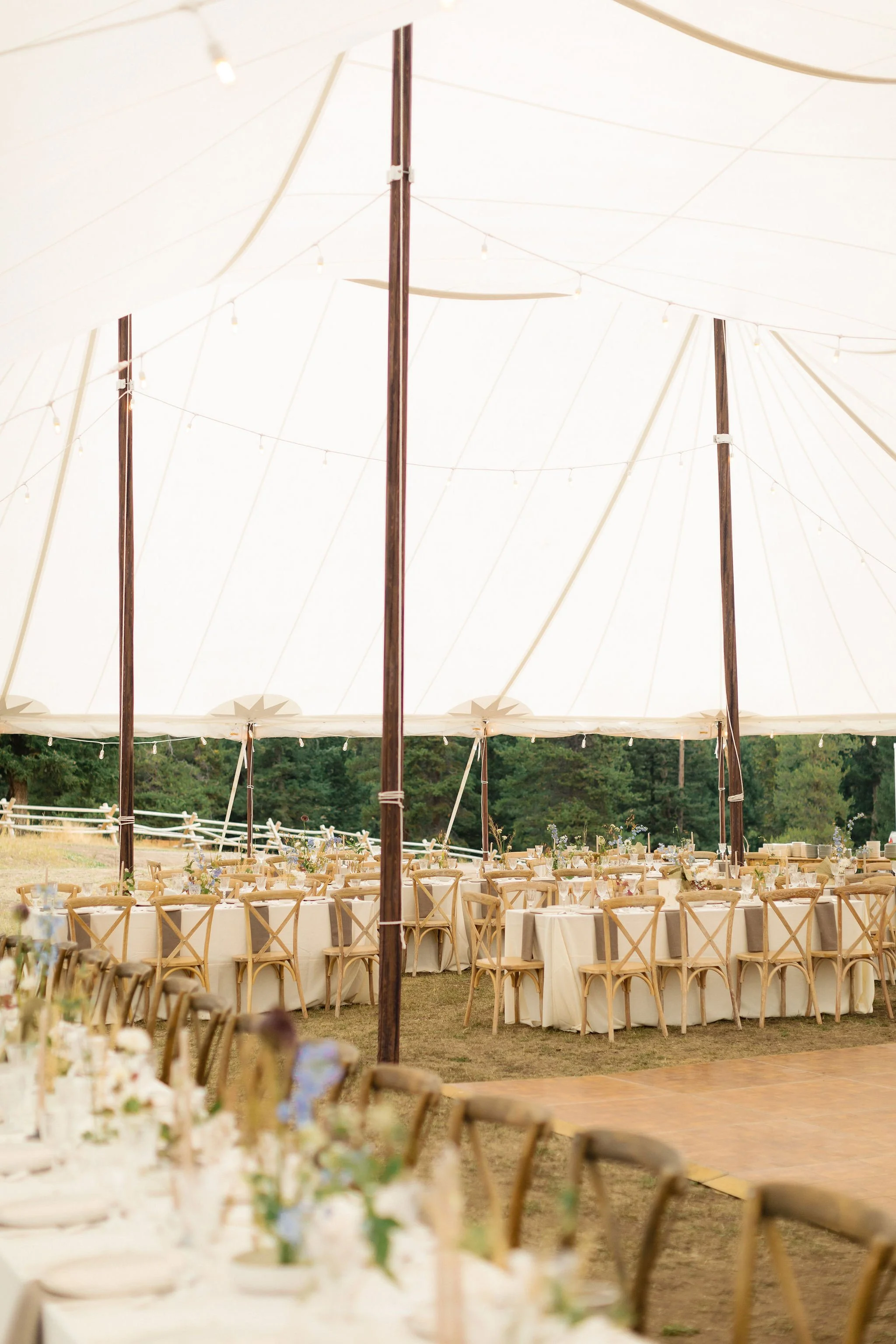 A large outdoor event tent with string lights, wooden poles, and round tables with cream tablecloths and chairs set for a celebration or gathering.