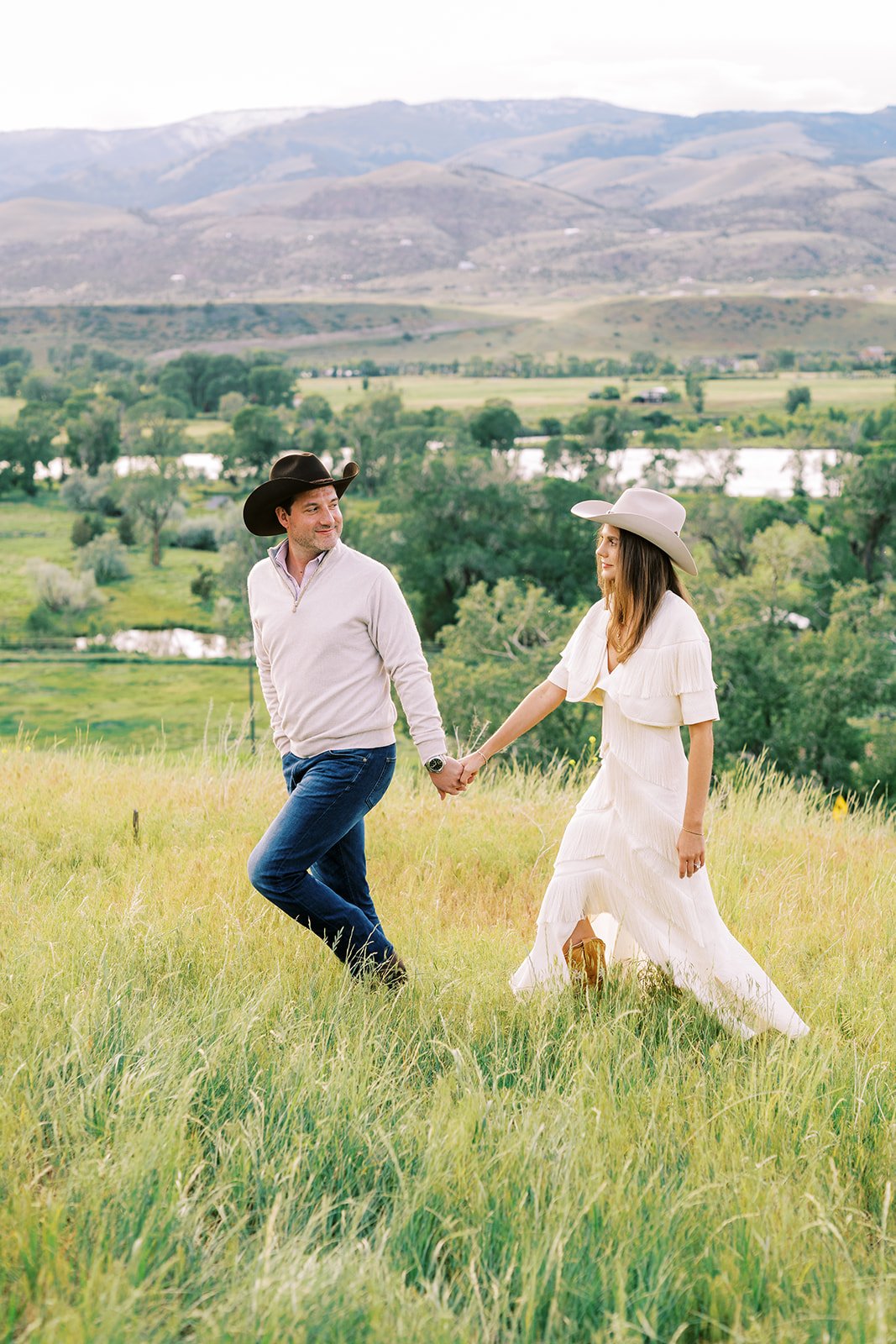 A couple holding hands and walking in a grassy field with a scenic background of rolling hills, trees, and a river.