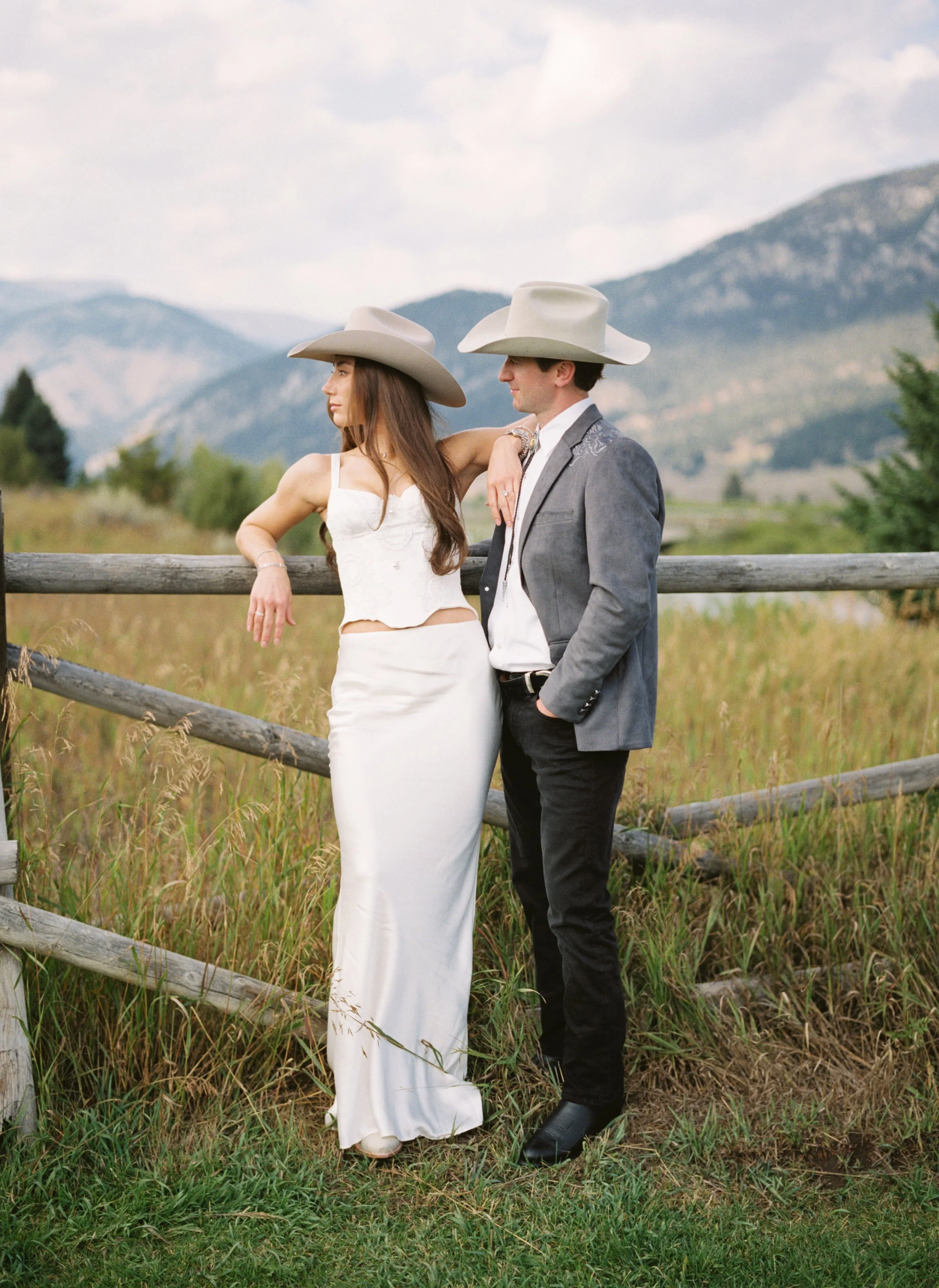 A woman and a man are standing next to a wooden fence outdoors. They are dressed in wedding attire, with the woman wearing a white dress and cowboy hat, and the man wearing a gray blazer, white shirt, and cowboy hat. Mountains and cloudy sky in the b