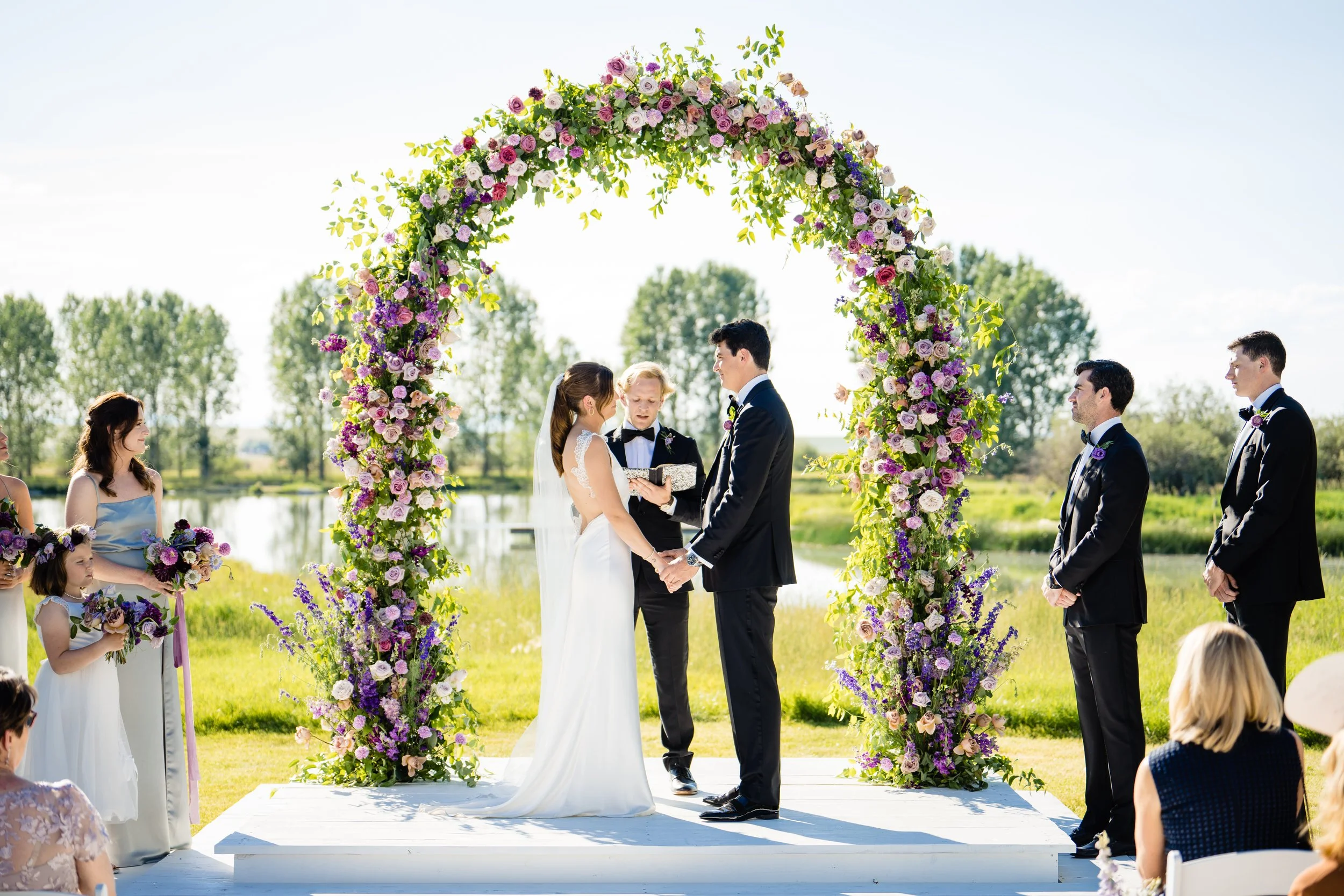 A wedding ceremony taking place outdoors with a couple exchanging vows under a floral arch, surrounded by family and friends.