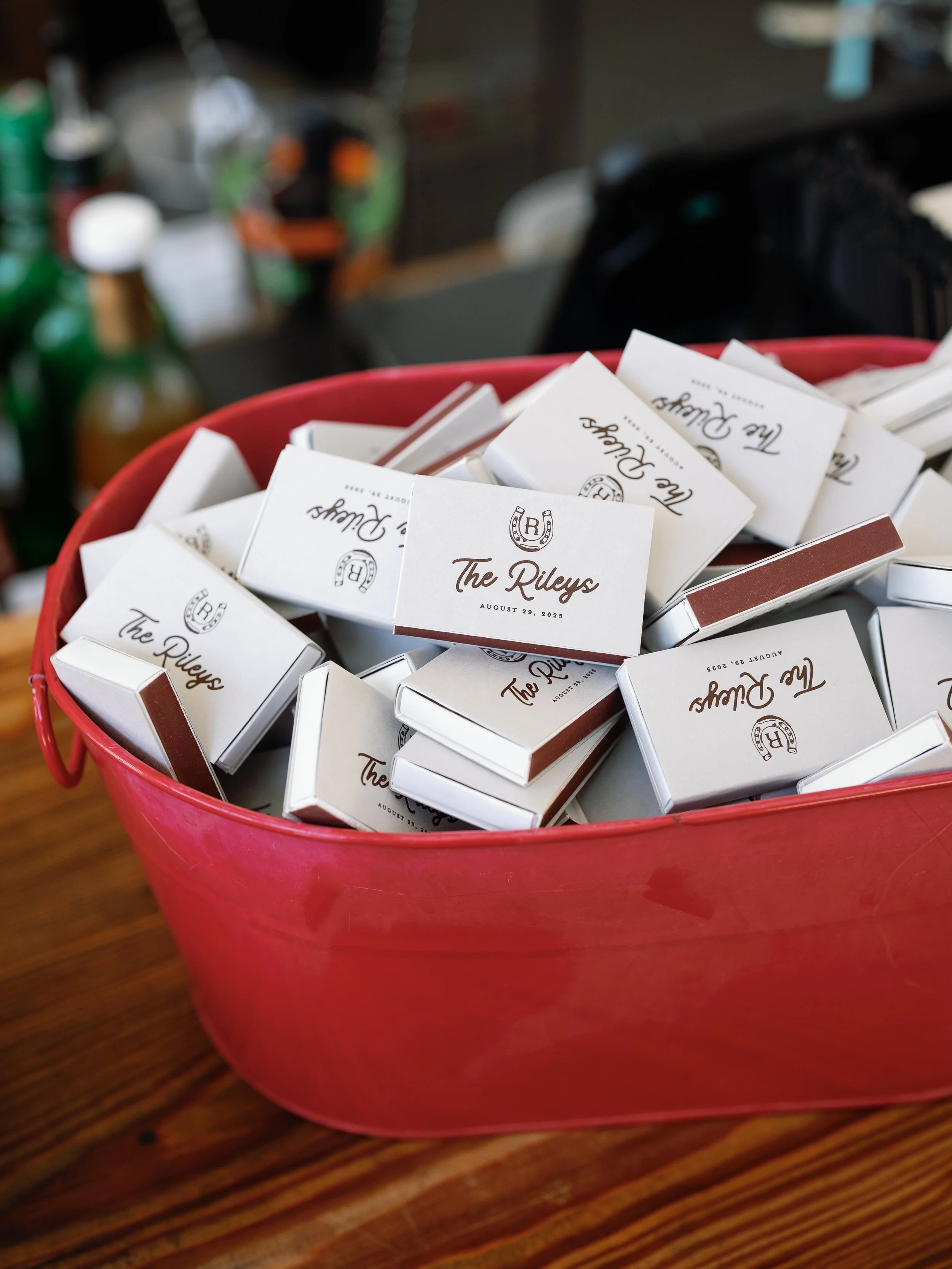 A red plastic basket filled with small white boxes labeled 'The Rileys' with a date of August 29, 2025, on the front, placed on a wooden surface.