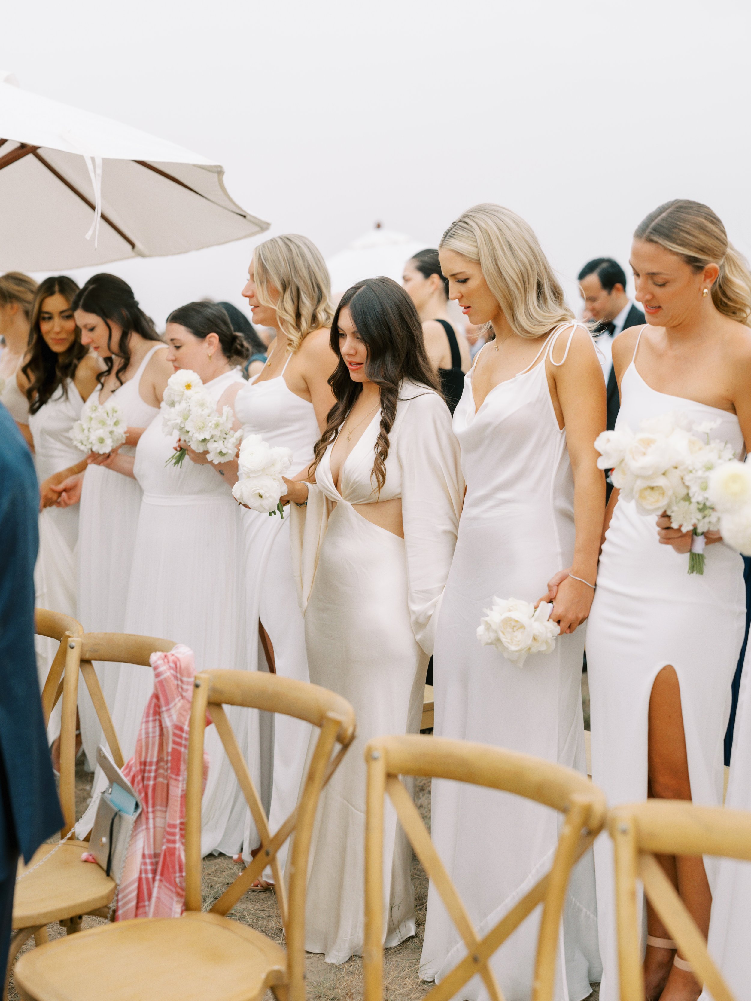 A group of women dressed in white, holding flower bouquets and standing together at an outdoor wedding ceremony.