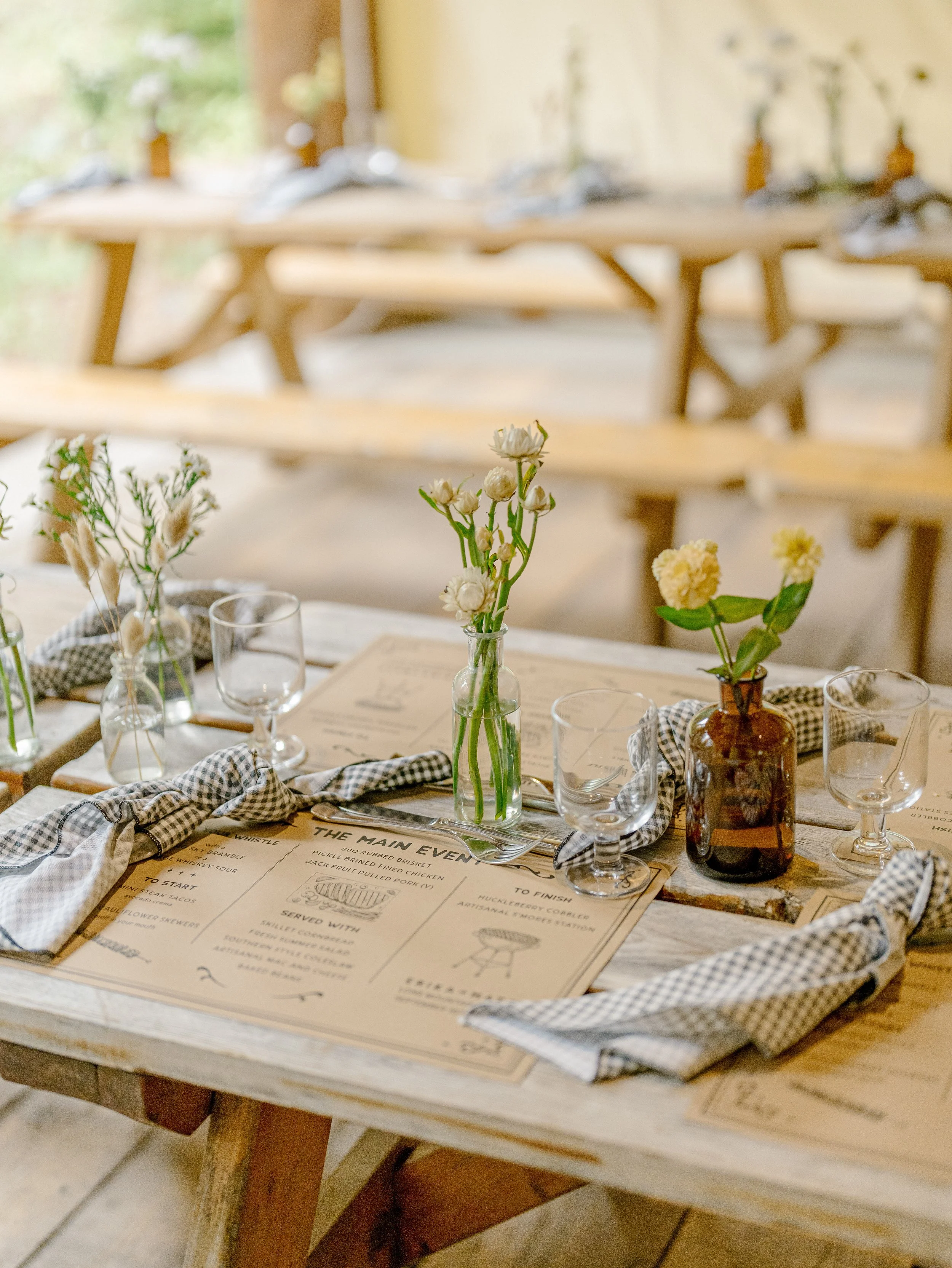 A table set with a floral centerpiece in a glass vase, glassware, cutlery, and a menu, with a rustic wooden bench and natural light in the background.