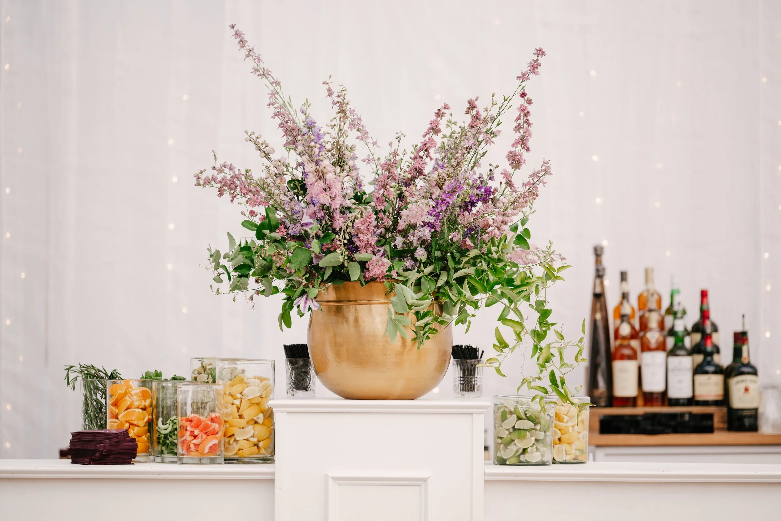 A large floral arrangement with pink and purple flowers in a beige vase sits on a white table. Behind the arrangement is a white wall with tiny string lights, and to the side, a bar area with various liquor bottles.