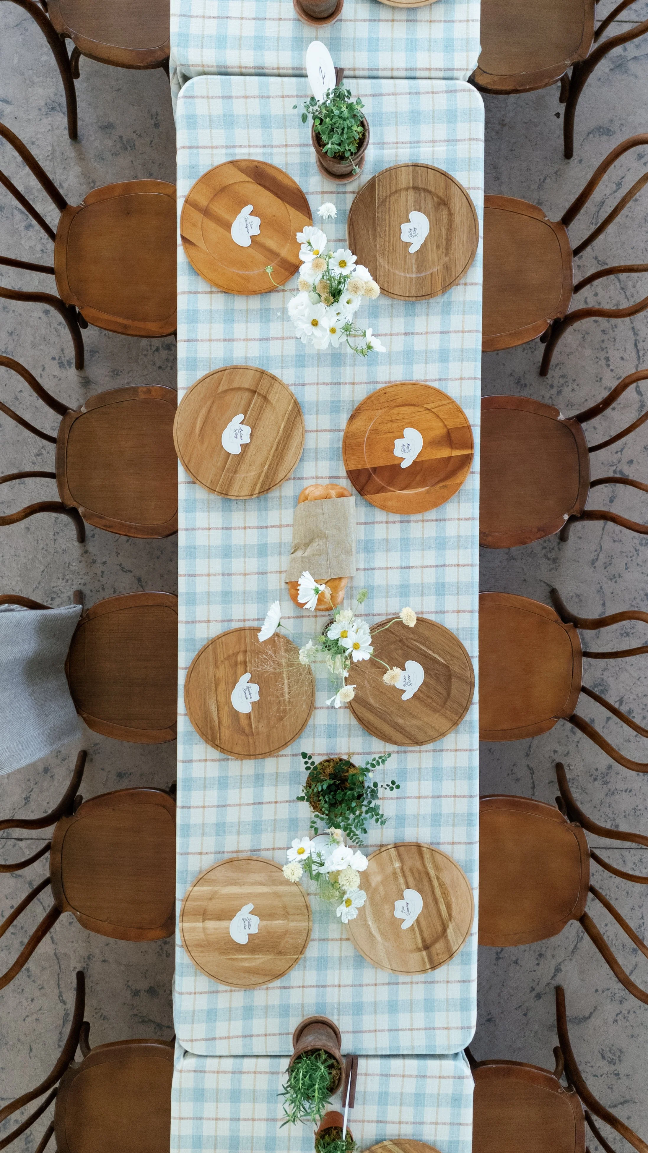 Top-down view of a long dining table with wooden chairs, decorated with a plaid tablecloth, white flowers, and small potted plants.