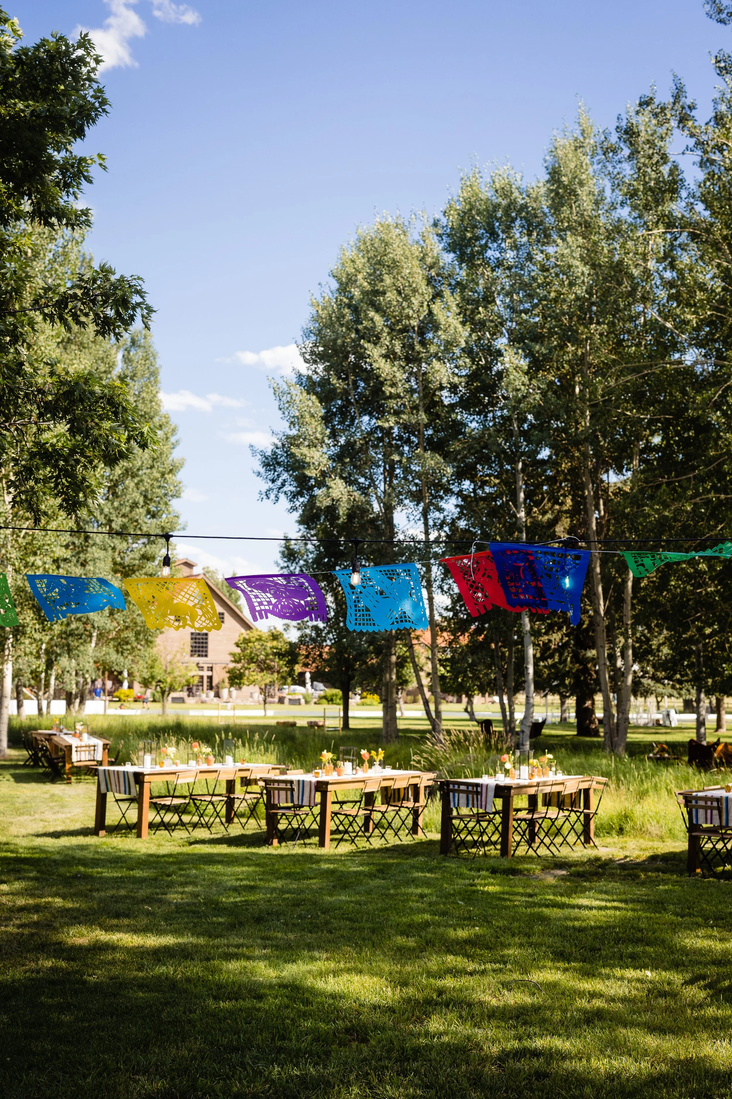 Outdoor celebration setup with long wooden tables, chairs, floral centerpieces, and colorful papel picado banners strung between trees in a lush green park.