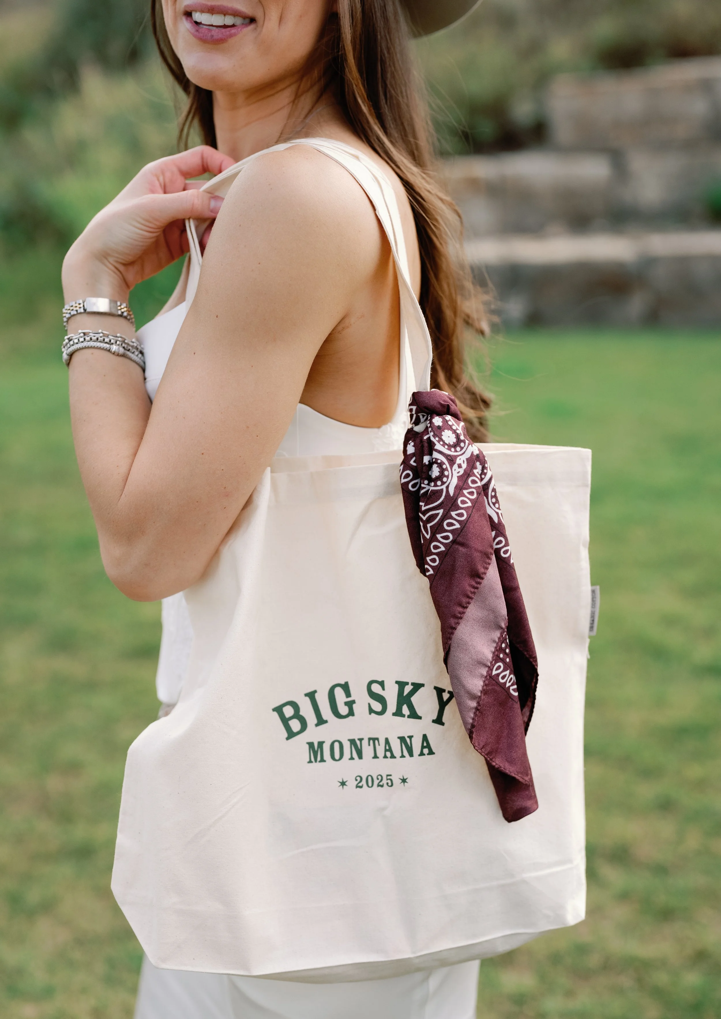 Close-up of a woman smiling, showing her teeth, with a tote bag in her hand. The tote bag has the words "Big Sky Montana 2025" on it and a maroon bandana tied to the handle. She is outdoors with a blurred green background.