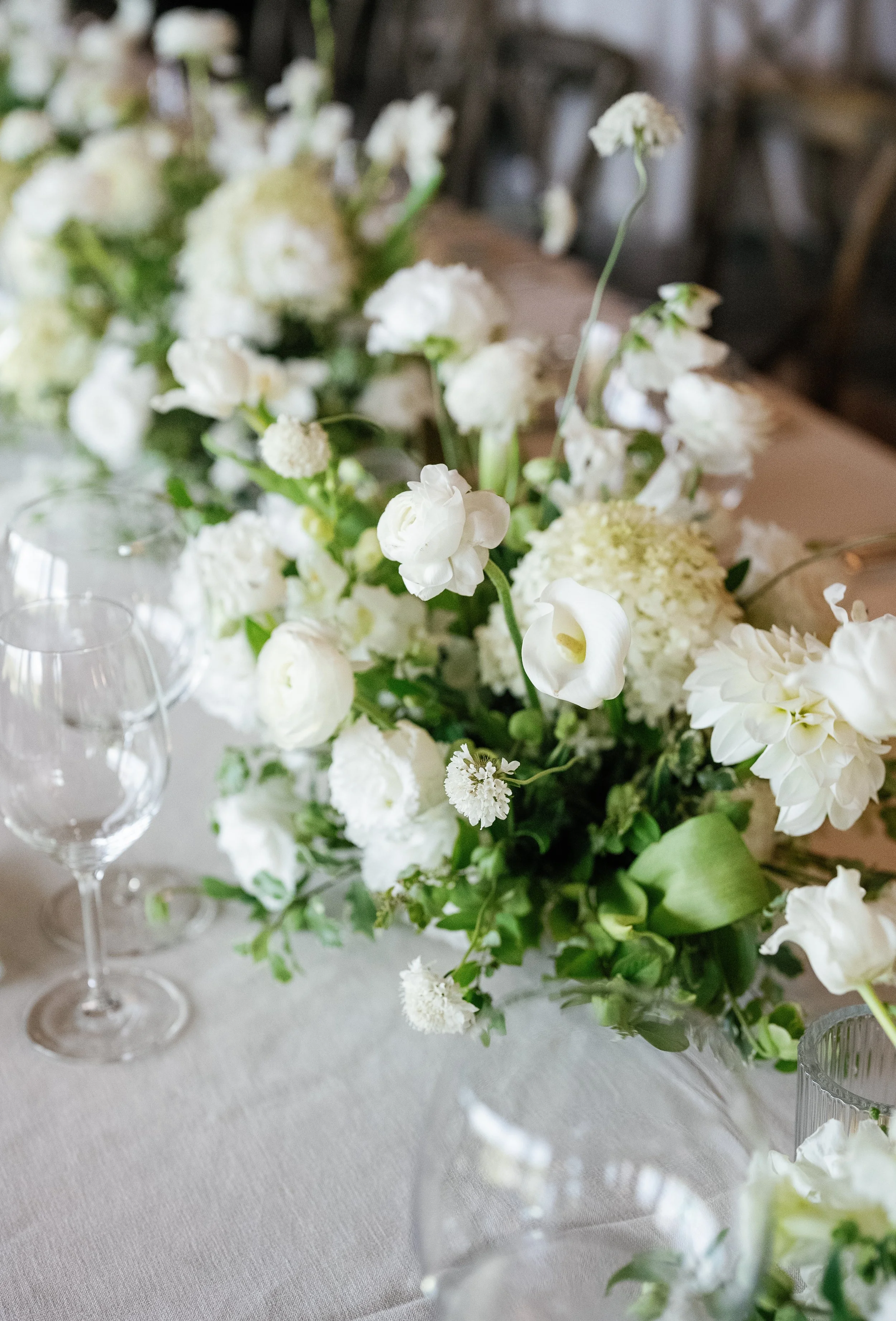A floral arrangement of white flowers on a table with glassware, set in a softly lit room.
