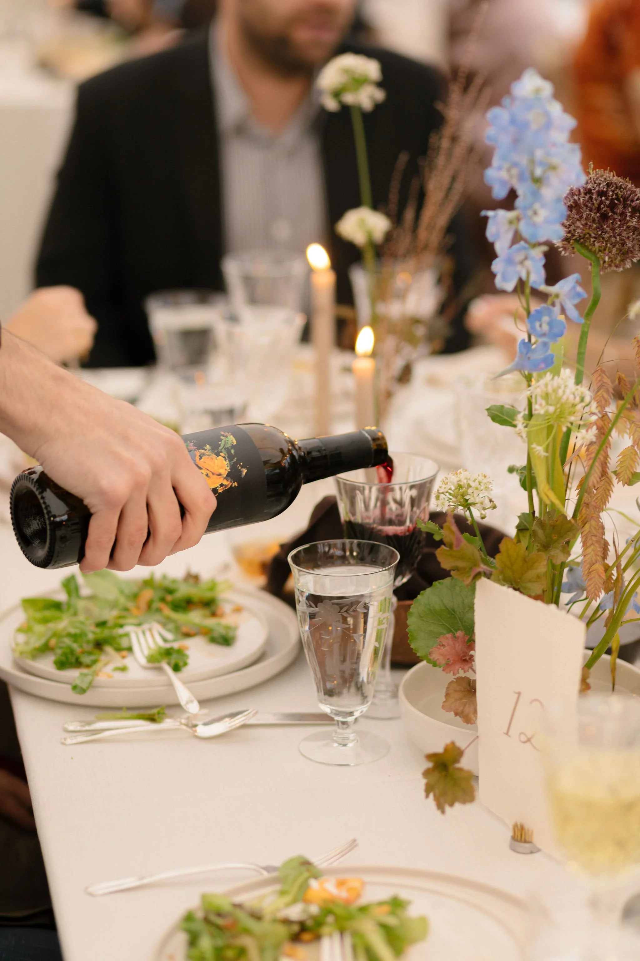 Person pouring red wine into a glass at a formal dining table decorated with flowers, candles, and plates with salad.