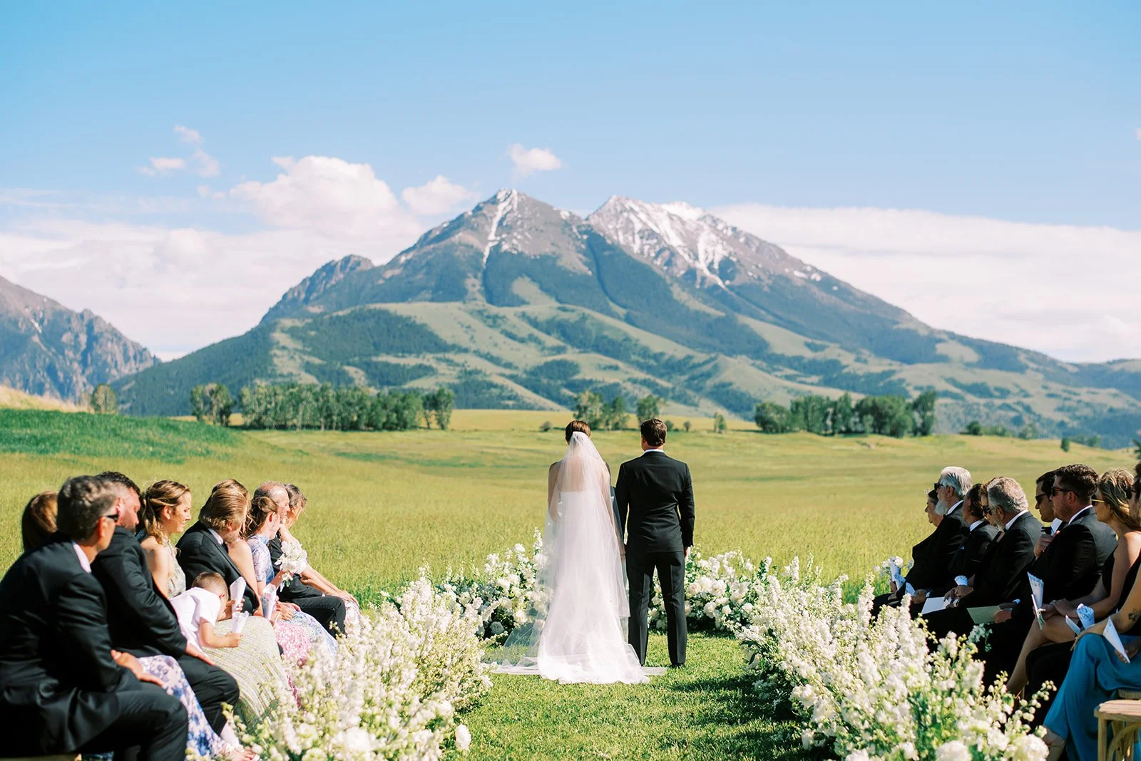 A wedding ceremony taking place outdoors in a green field with mountains and a blue sky in the background. The bride and groom are standing together with their backs to the camera, facing the officiant. Guests are seated on either side of the aisle, 