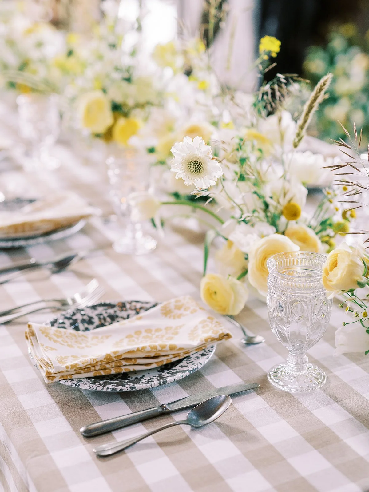 A beautifully set dining table with a floral centerpiece of white and yellow flowers, crystal glasses, patterned plates, gold-colored napkins, and silver cutlery on a checkered tablecloth.