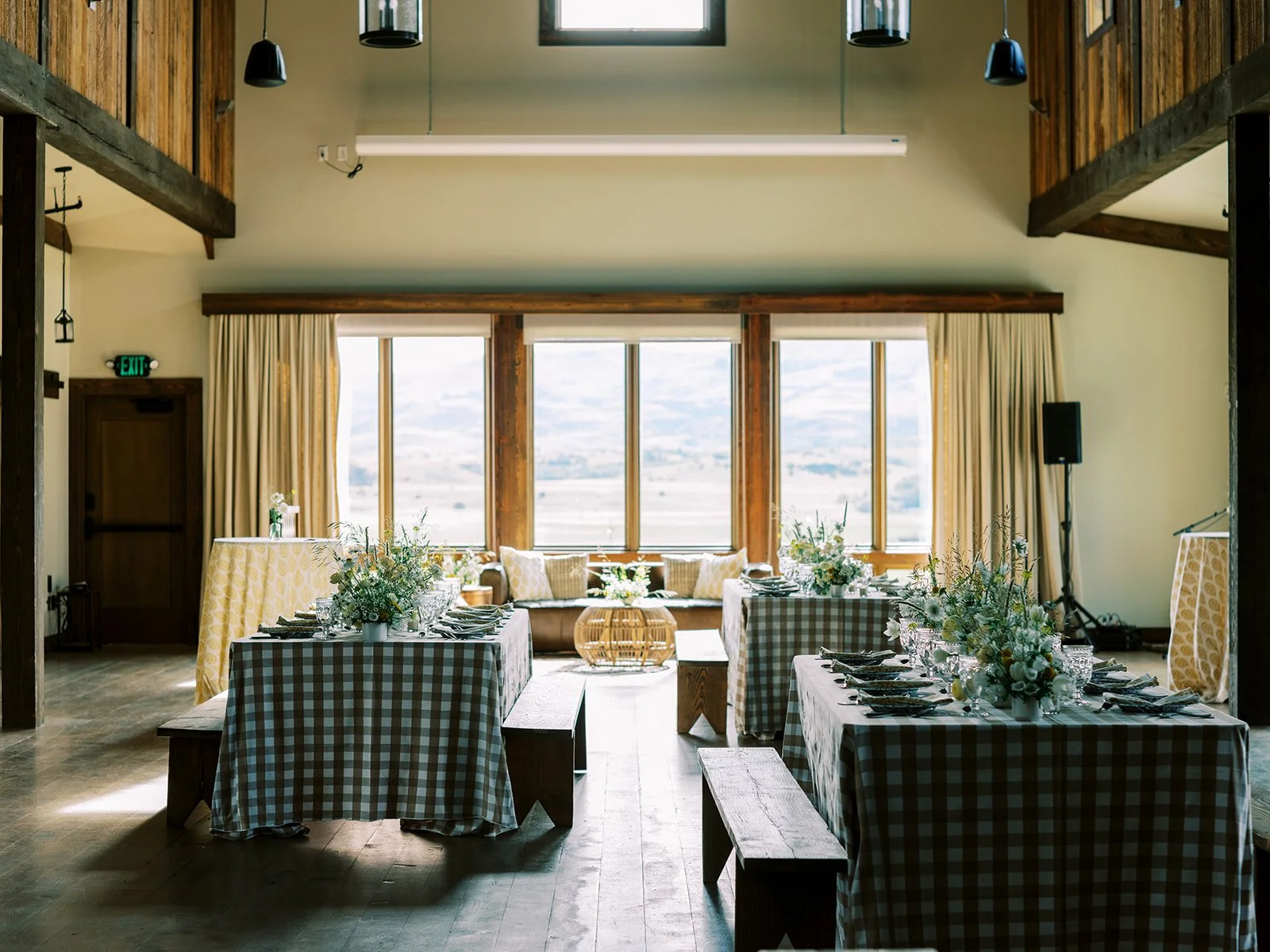 Indoor dining area with long tables covered in checkered tablecloths, floral centerpieces, and place settings. Large windows with curtains provide natural light, and there is a cozy window seat with cushions. Rustic wood beams and modern black pendan