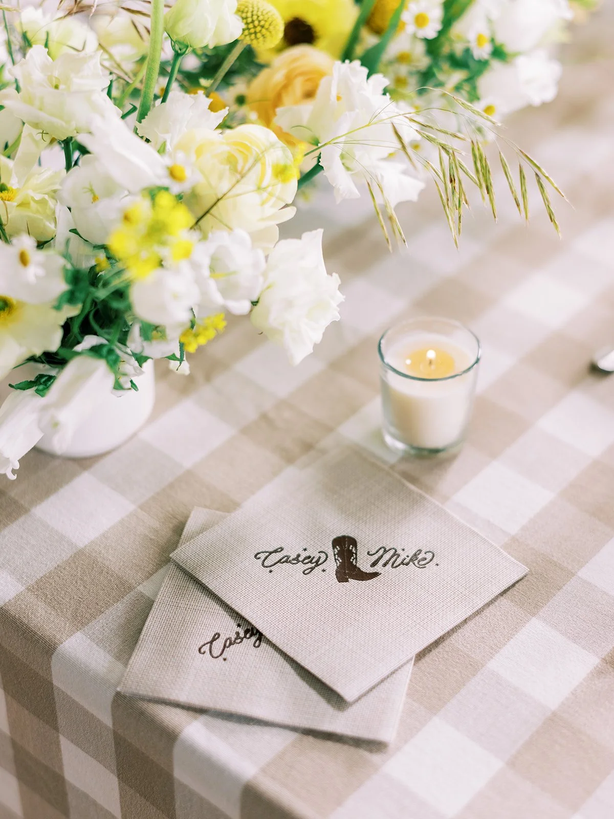 A table setting featuring a floral centerpiece with white and yellow flowers, a lit candle in a glass holder, and a card with the names "Taisy" and "Mike" written on it.
