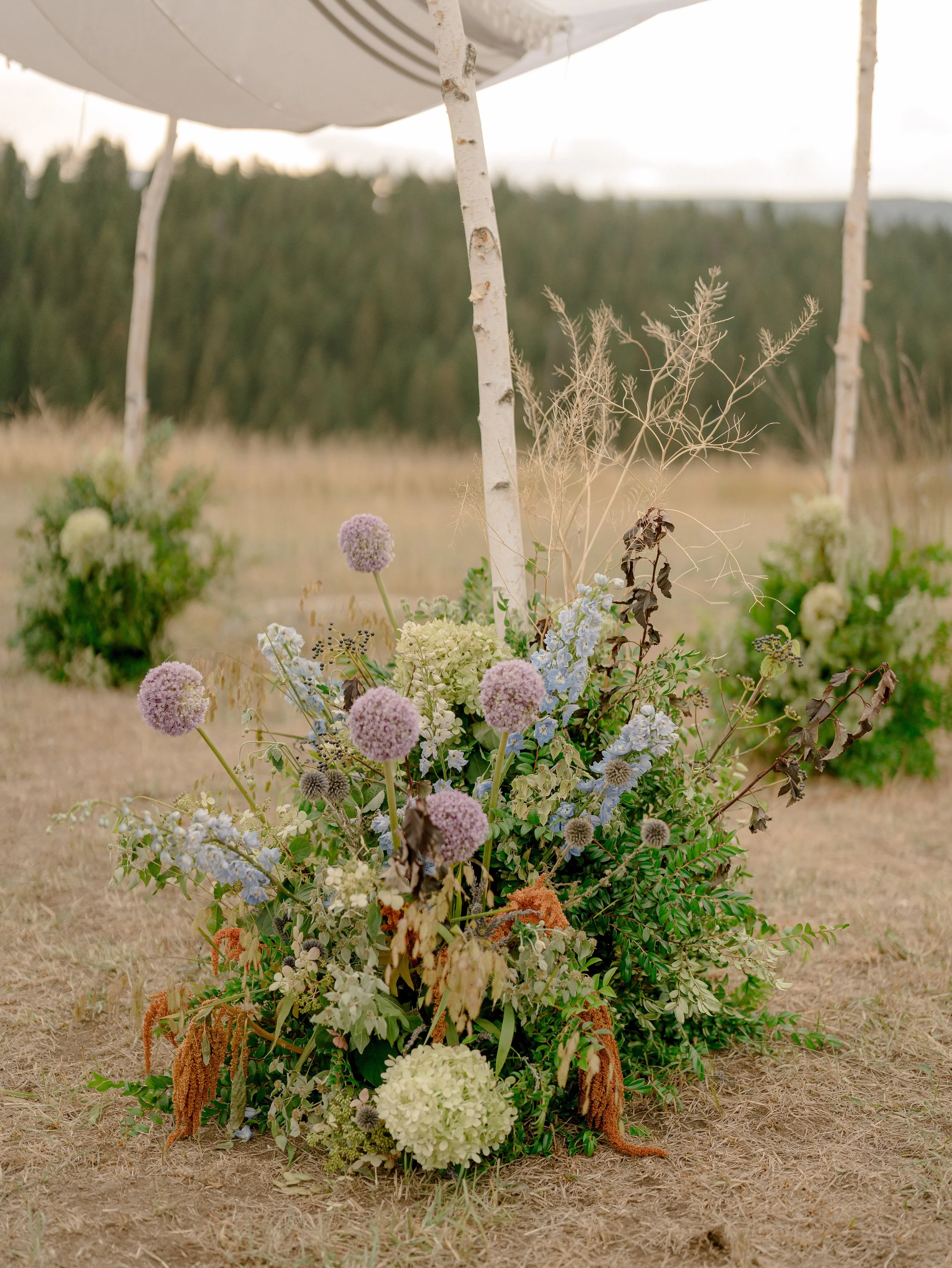 A floral arrangement with white, pink, purple, and green flowers, accented with dried plants, positioned outdoors on dry grass, with a blurred background of trees and an overcast sky.