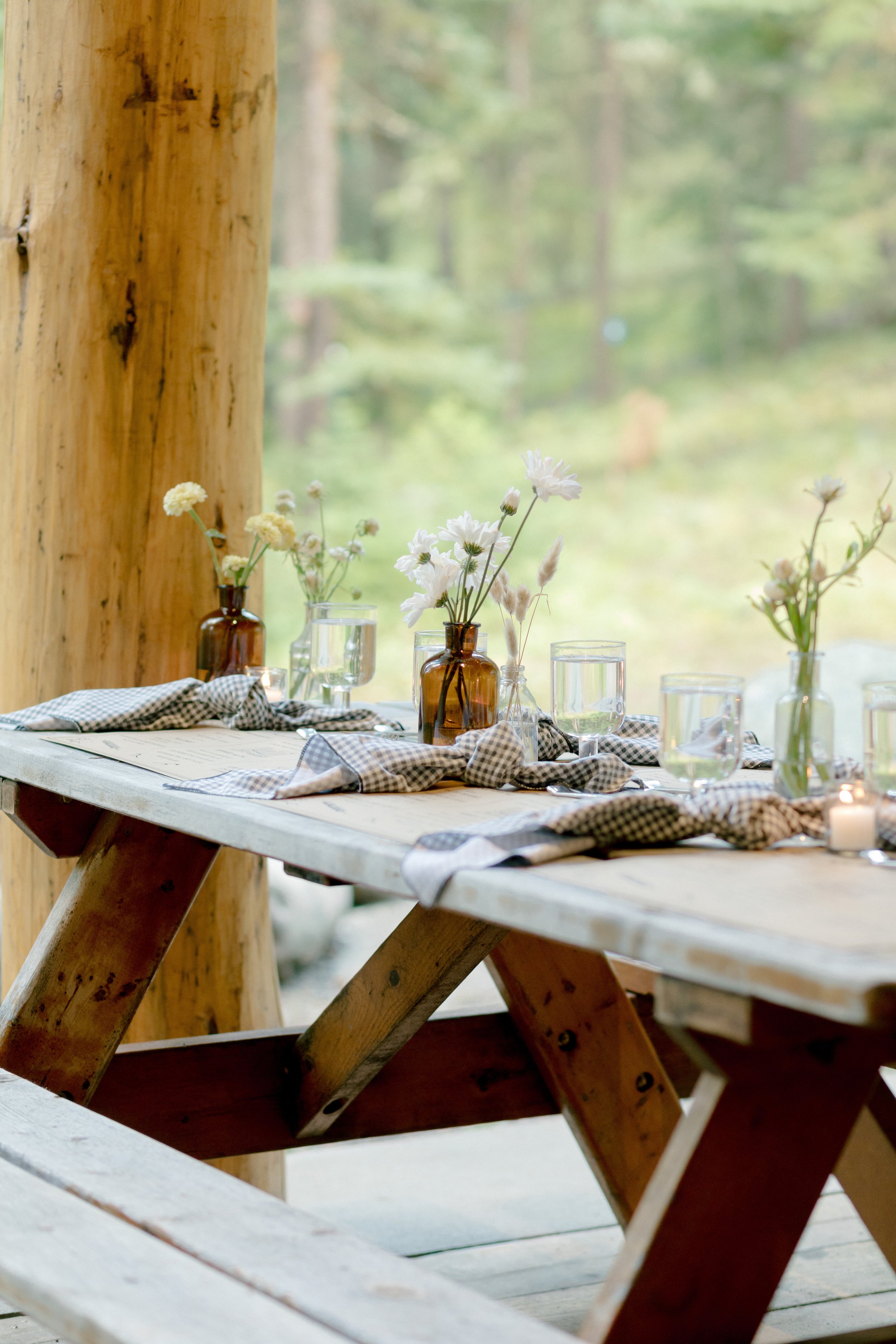 A rustic wooden table set with glass bottles containing white flowers, surrounded by checkered cloths, in front of a large window with a forest background.
