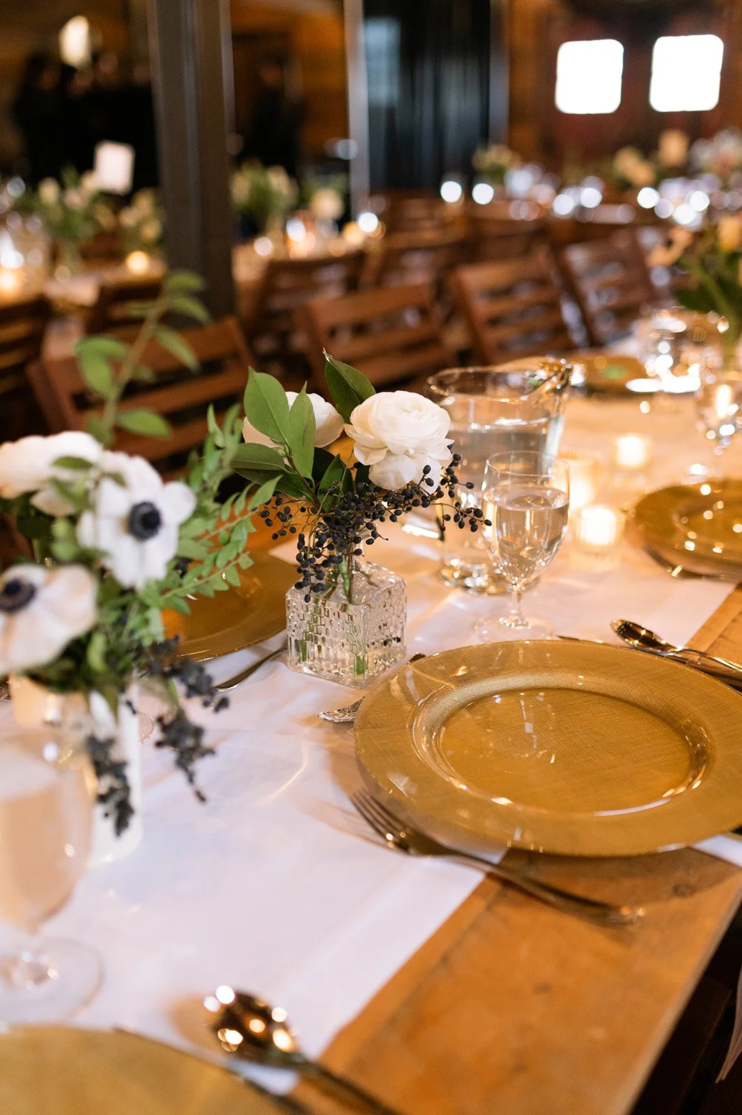 Warm rustic wedding reception tablescape with gold charger plates, white floral centerpieces, and candlelight, designed by Wild Blume