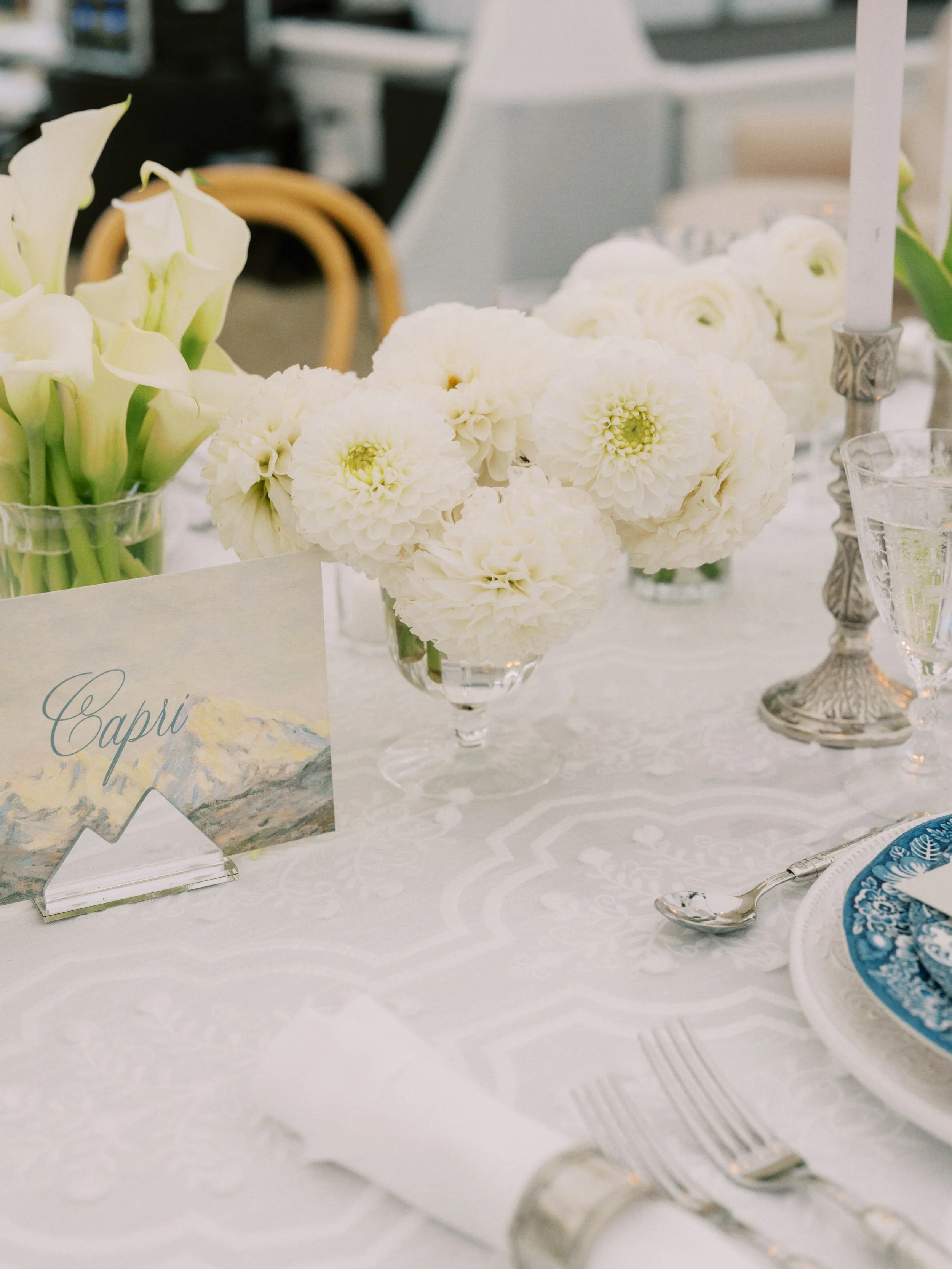 A centerpiece of large white flowers on a table set for an event, with silver candlesticks, glassware, and a blue and white patterned plate.