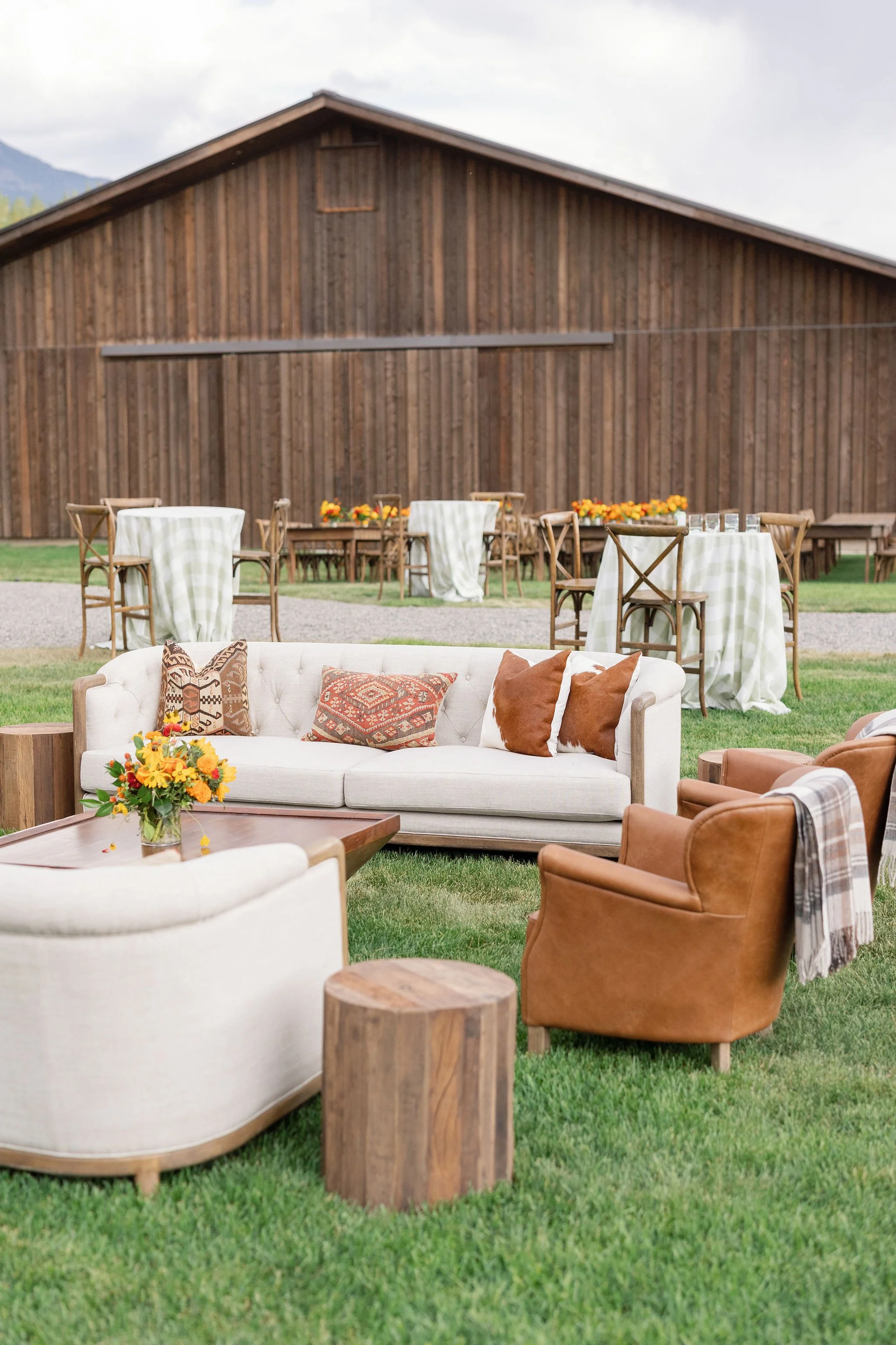 Outdoor wedding lounge setup with white sofas, leather chairs, and wood stump accents beside a rustic barn, designed by Wild Blume