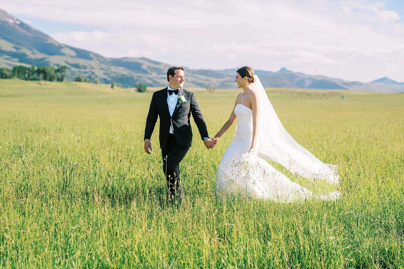 A bride and groom holding hands and walking through a grassy field on their wedding day, with mountains and a partly cloudy sky in the background.