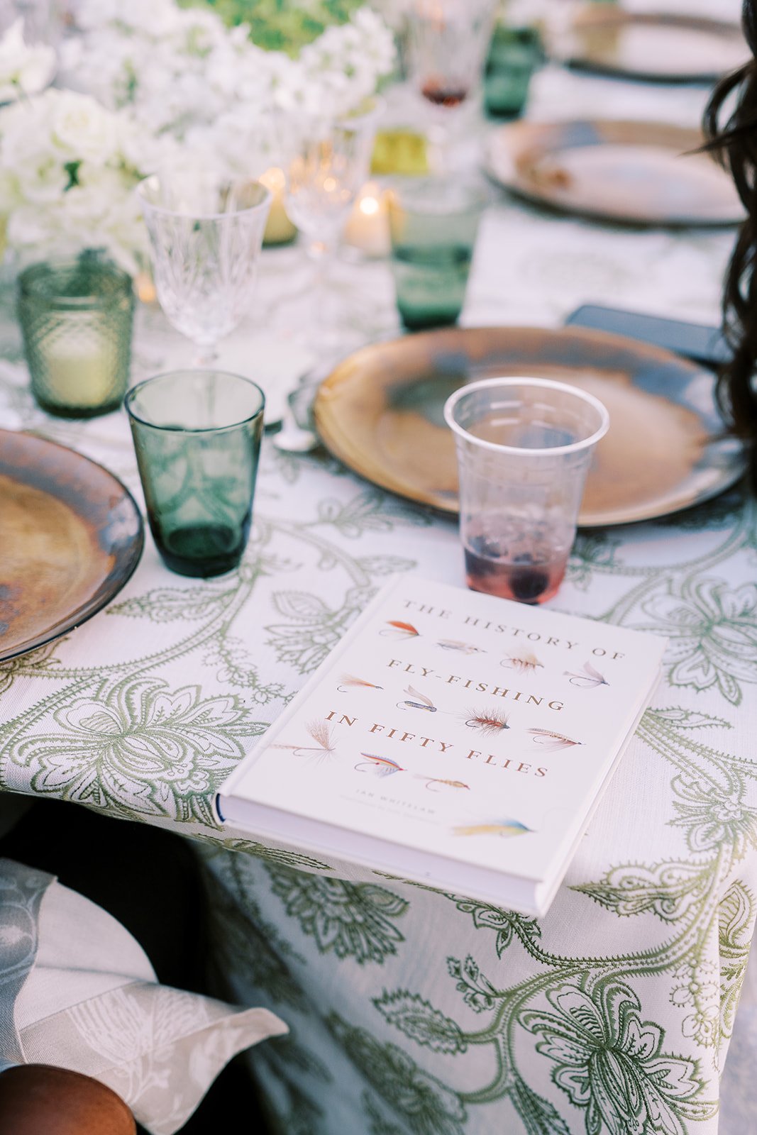 A table with a floral tablecloth, glassware, plates, and a book titled 'The History of Fly-Fishing in Fifty Flies'.