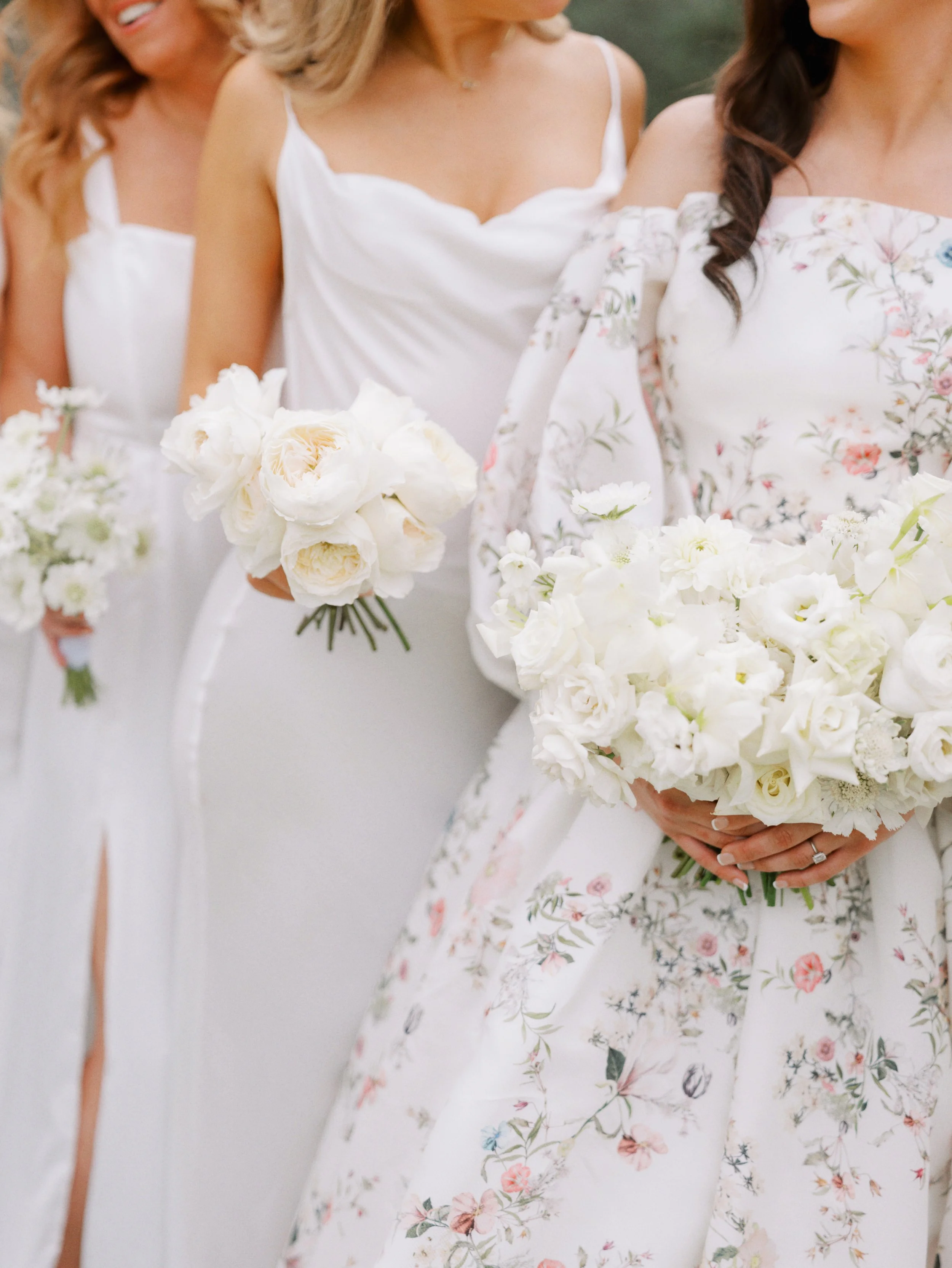 Three women dressed in white and floral dresses holding bouquets of white flowers at an outdoor wedding or event.