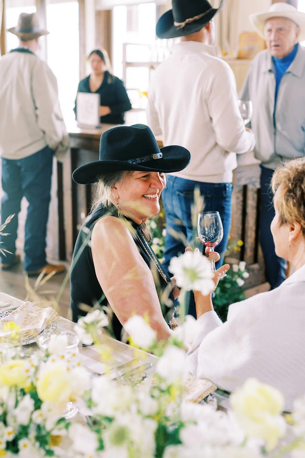 People at a social gathering, with some wearing cowboy hats, engaging in conversation, with floral decorations and wine glasses on the table.