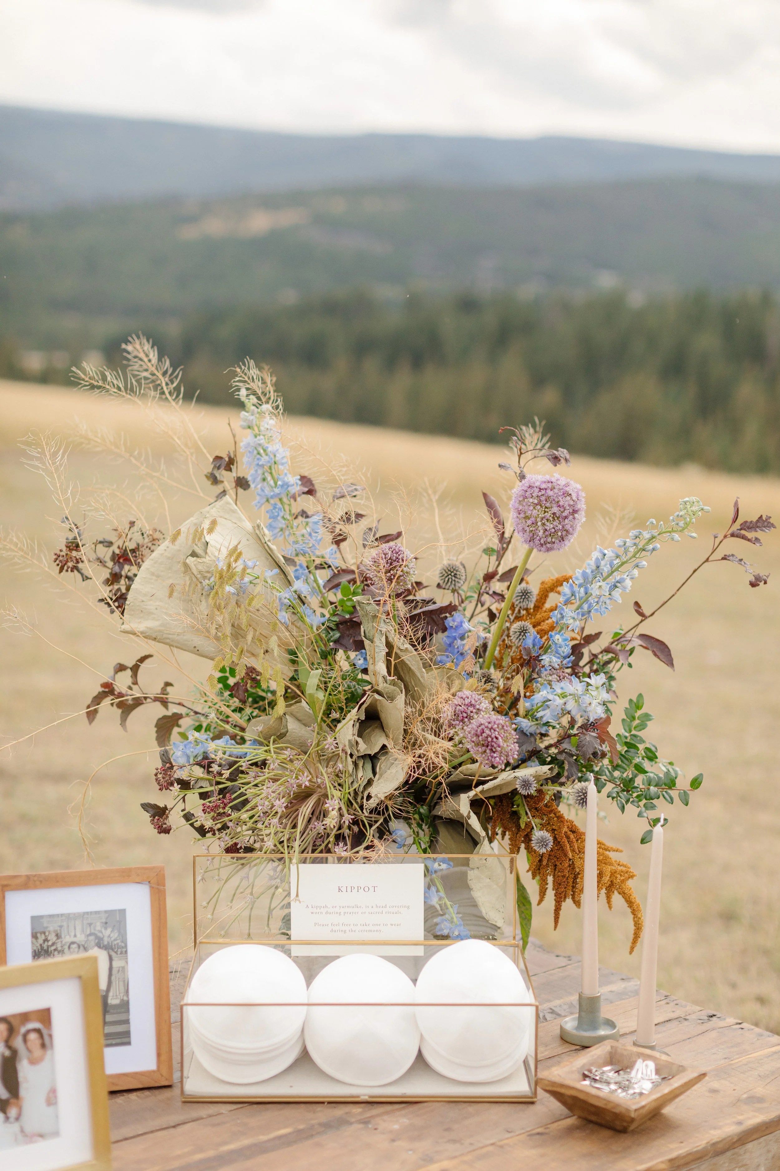 A rustic outdoor wedding display with a bouquet of mixed wildflowers in front of a scenic mountain landscape. Includes framed photographs, standing candles, and a sign labeled 'Kippot'.