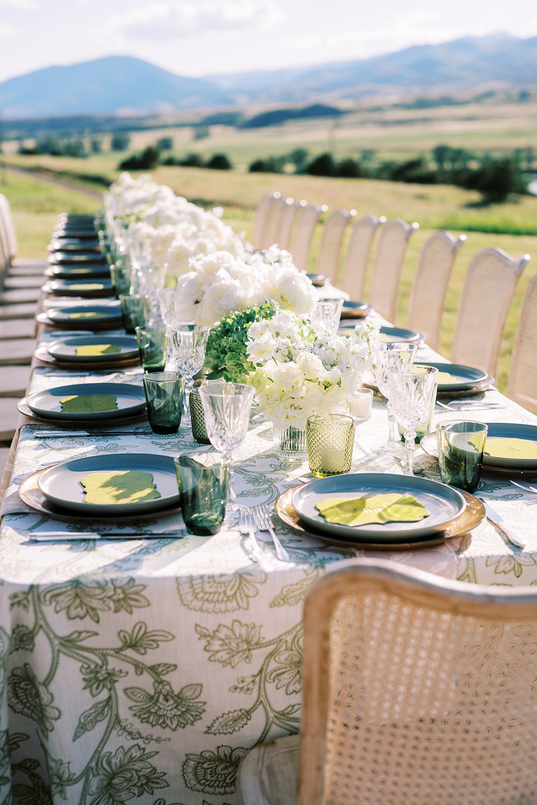 Outdoor table set for a meal with white floral centerpieces, plates, glasses, and silverware, against a background of rolling hills and mountains.