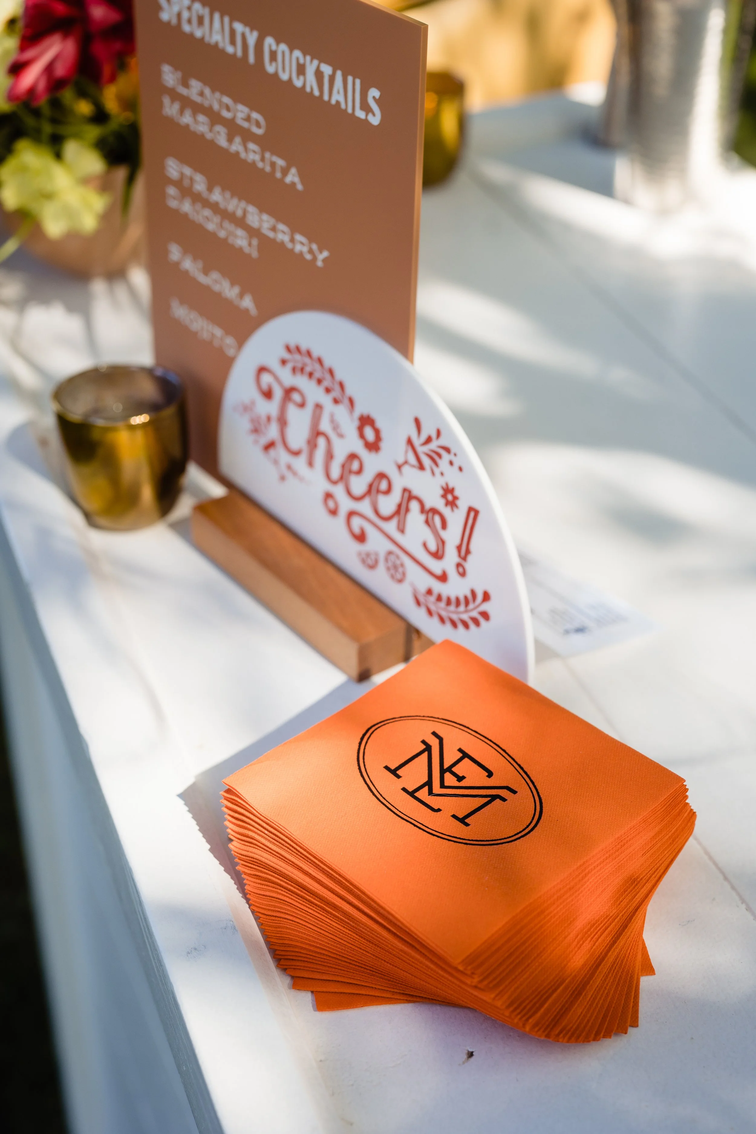 Close-up of a menu sign listing specialty cocktails, a gold shot glass, and orange napkins with a logo on a white table at an outdoor event.