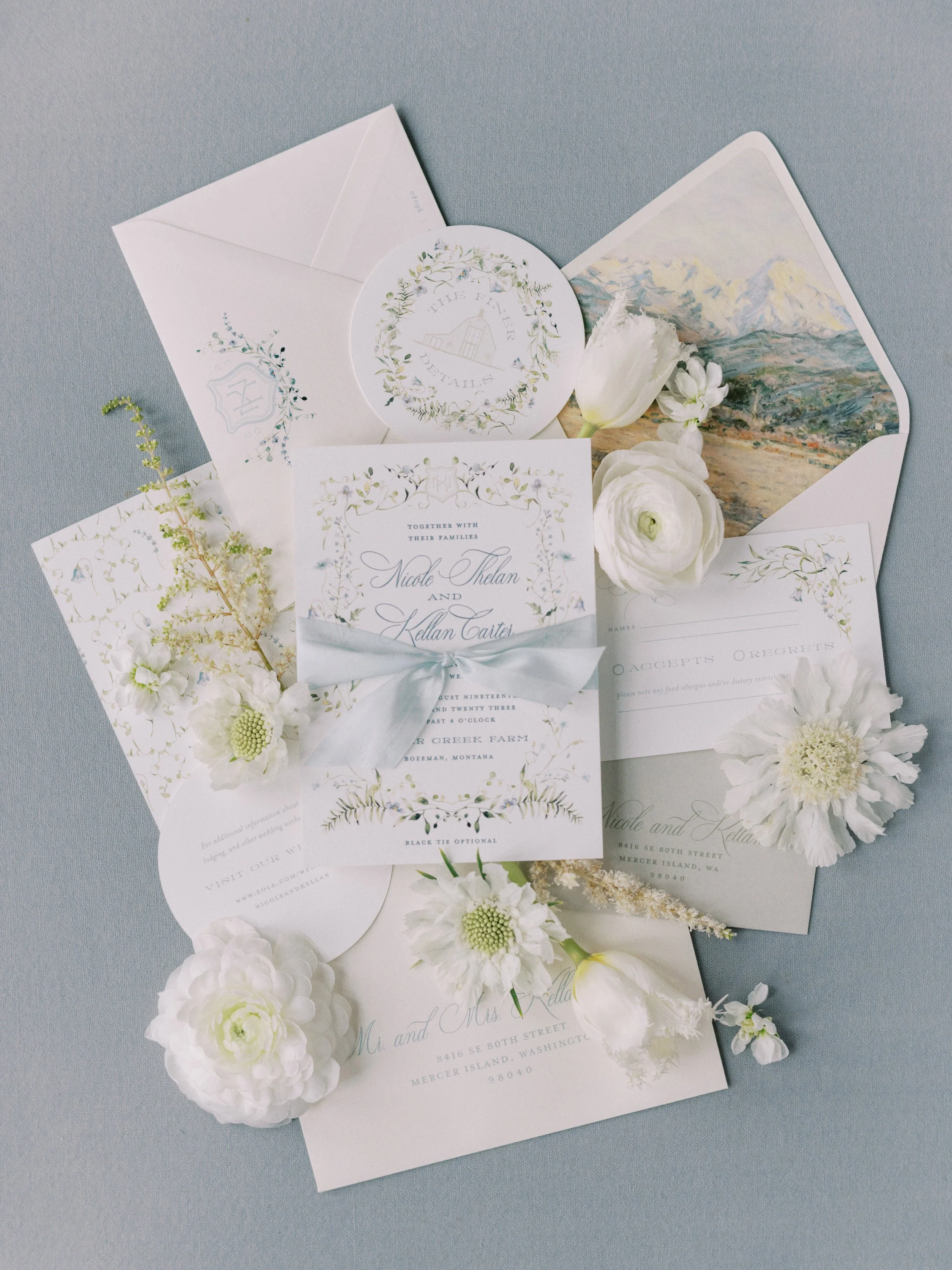 Collection of wedding stationery including invitations, RSVP cards, and envelope, decorated with white flowers and greenery, with a landscape painting in the background.