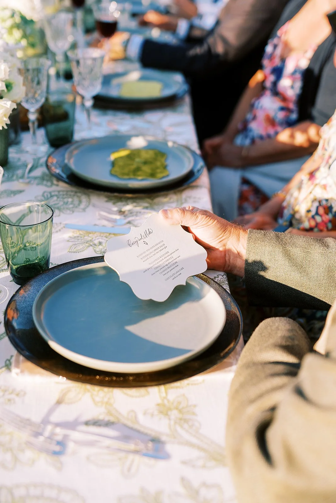 People seated at a decorated outdoor dining table with plates, glasses, and a menu. One person is holding a menu with a scalloped edge.